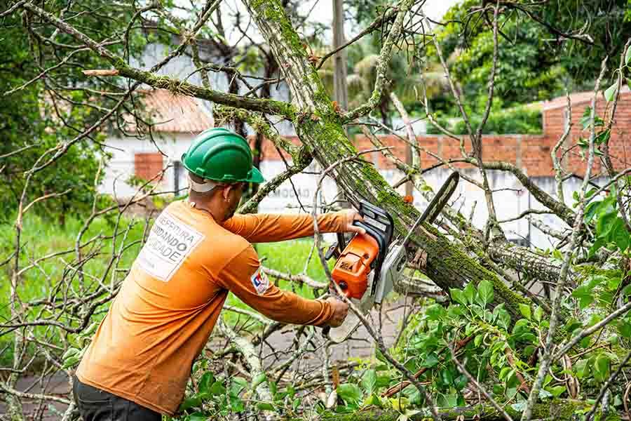 A person in a green hard hat and orange shirt uses a chainsaw to cut a fallen tree branch outdoors.