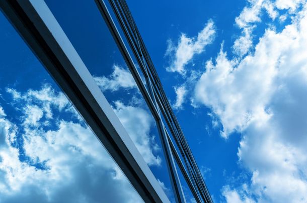 Fachada de vidro arquitetônico refletindo céu azul com nuvens, ideal para aplicação de película arquitetônica que mantenha transparência e reduza calor.