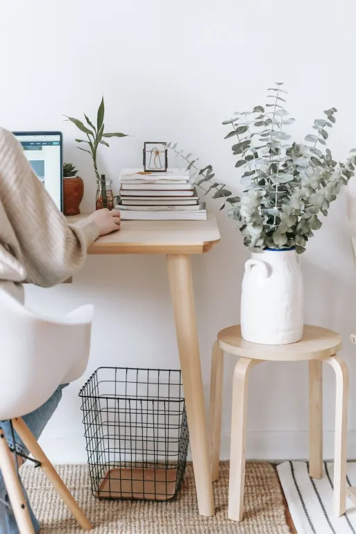 Home desk with laptop, stacked books, potted plants, and a white vase on a wooden stool by a wicker basket.