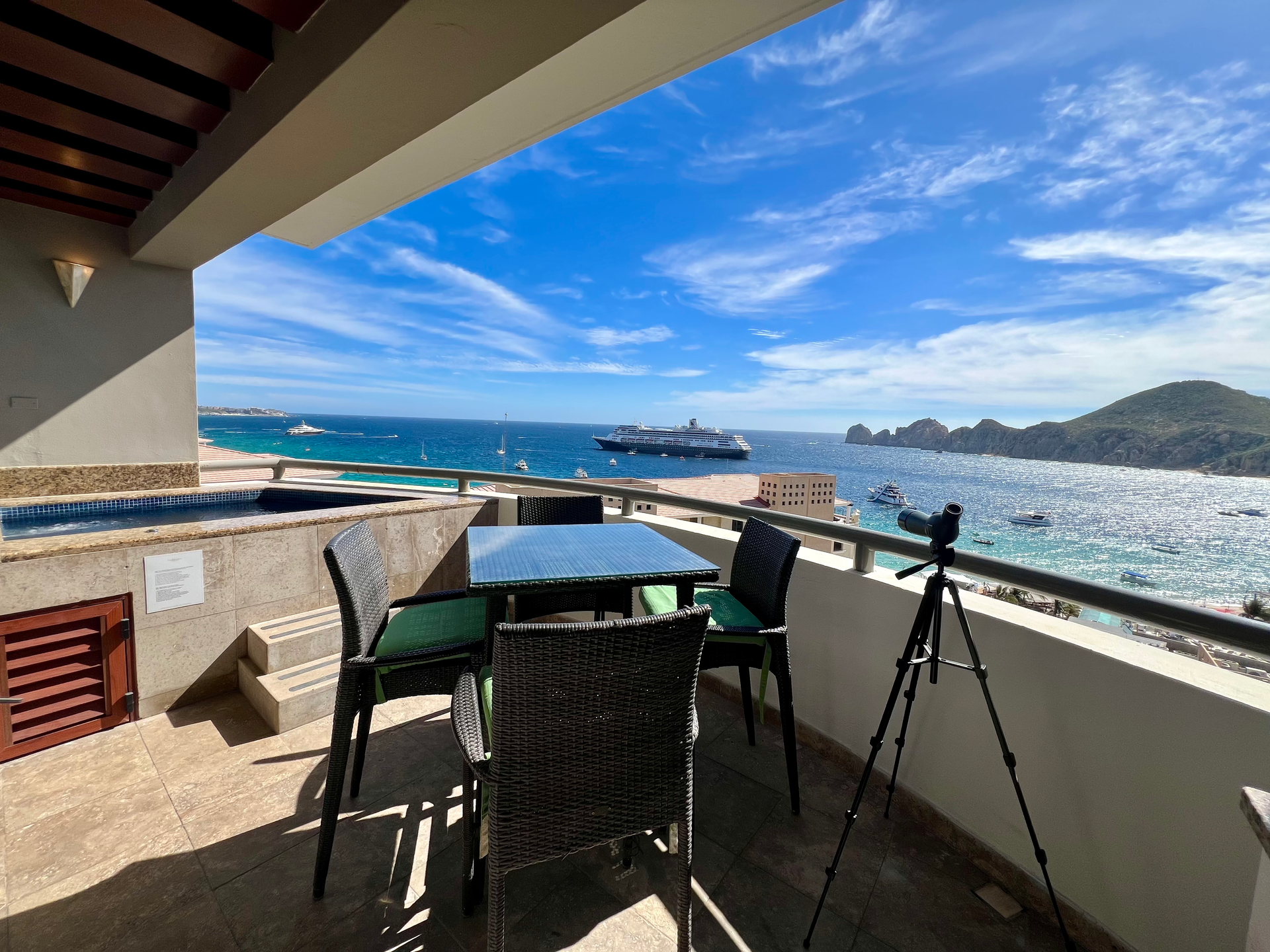 Balcony overlooking a blue ocean bay with a table, chairs, and a tripod beside the railing.