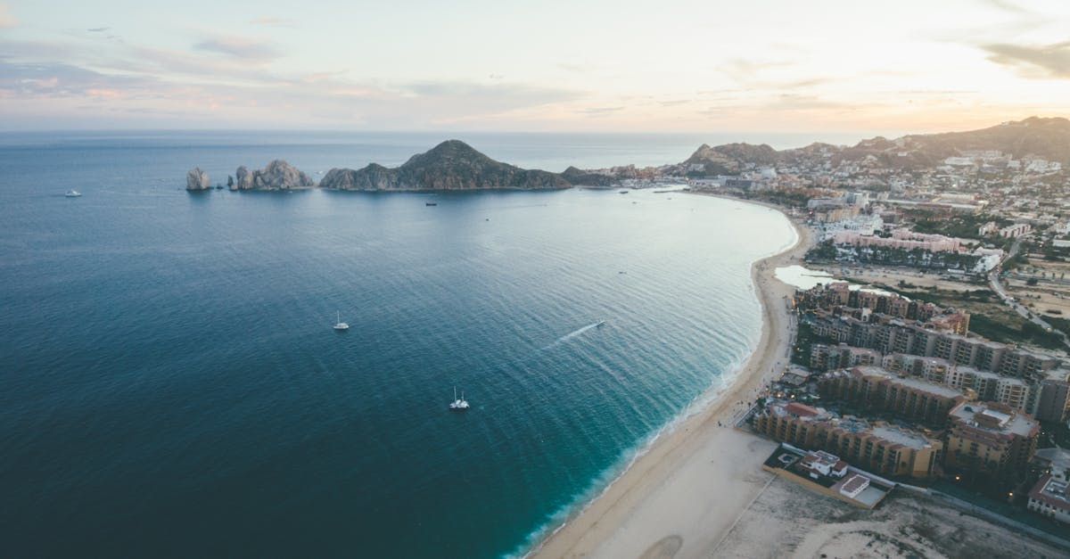 Aerial view of a curving beach and calm blue bay beside a coastal city with buildings and small islands.
