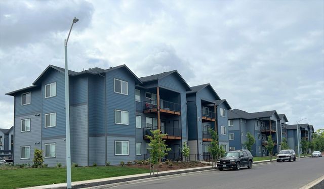 A row of blue apartment buildings with cars parked in front of them on a cloudy day.
