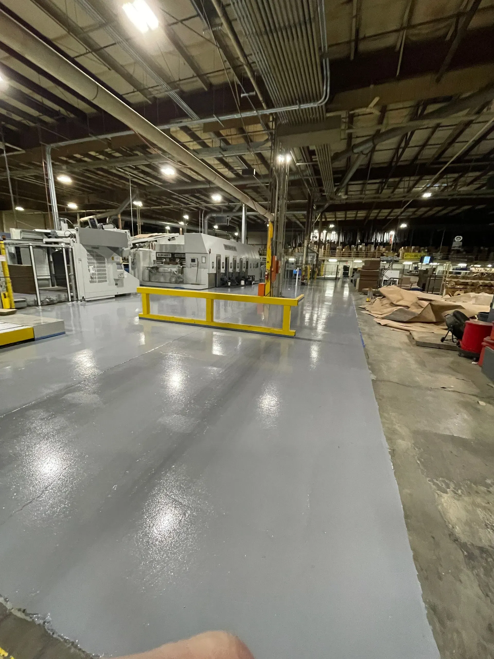 A wide-angle view of a factory floor featuring a newly applied, glossy light gray epoxy floor and a yellow safety guard.