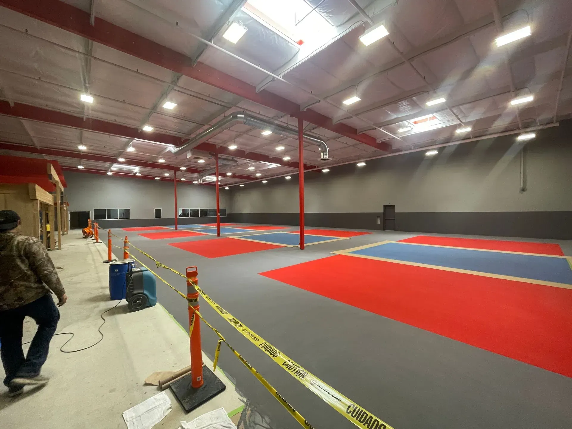 Large warehouse interior with red, blue, and grey mats. A person walks near caution tape, overhead lights.