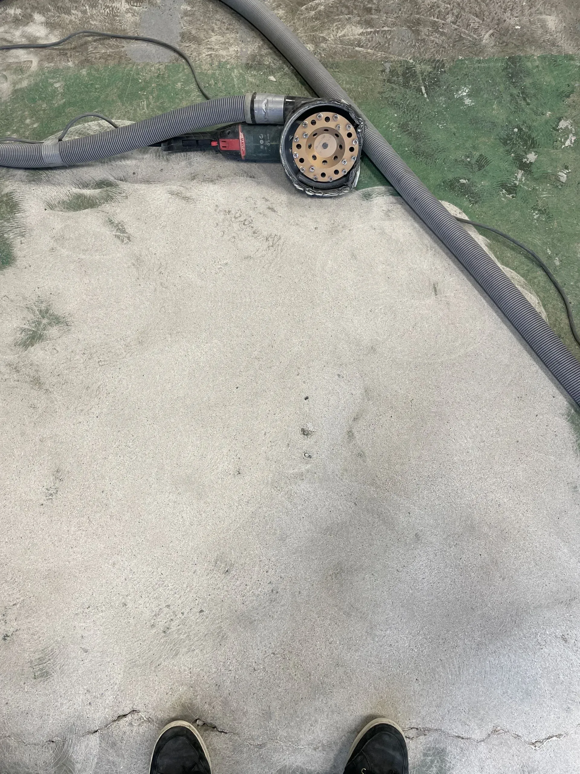 Foot-level view of a concrete floor with a sanding machine and hose. Black shoes are at the bottom.