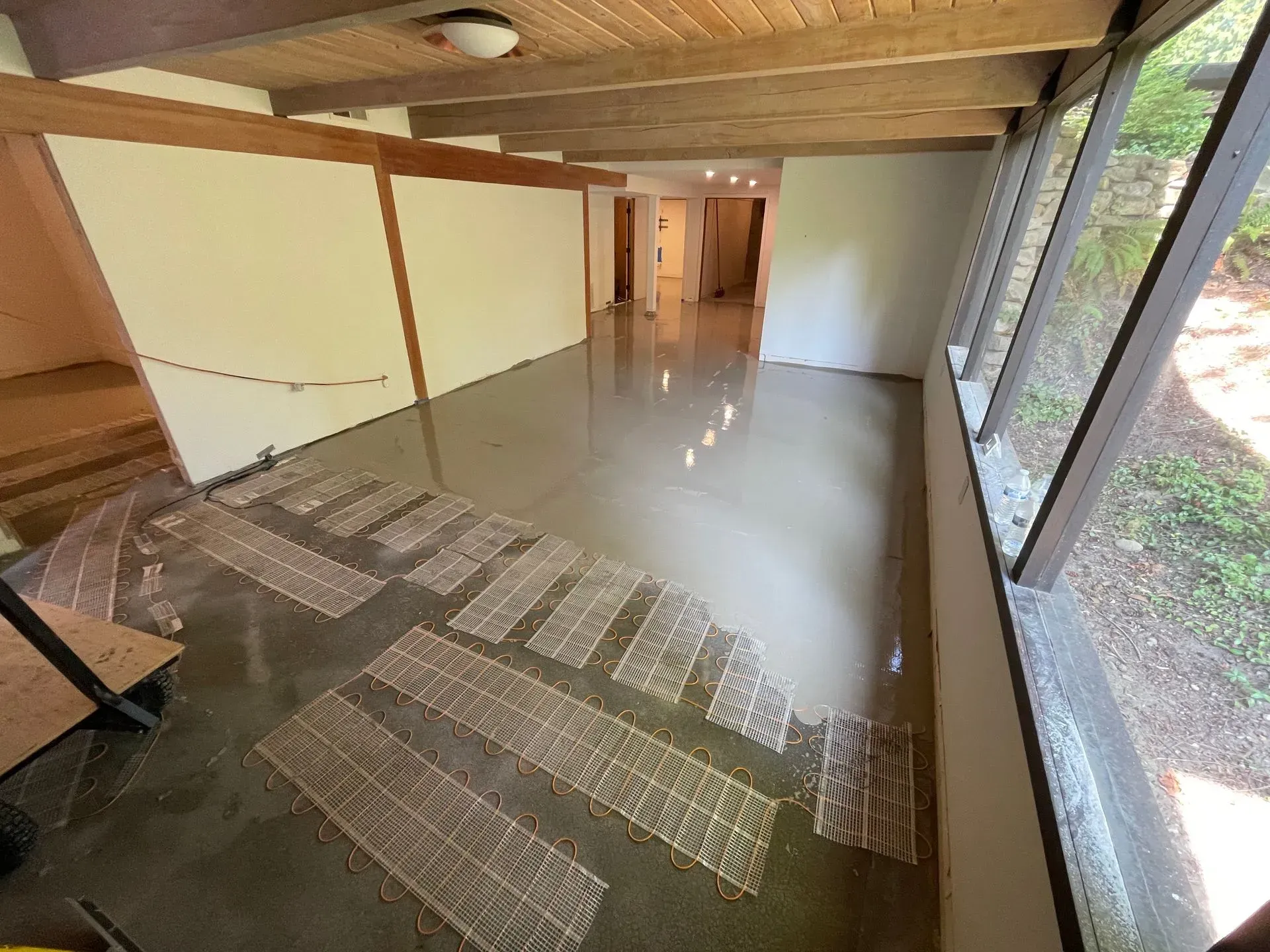 Interior view of a room with exposed beams, polished concrete floor, and large windows. Wood panels are laid on the floor.