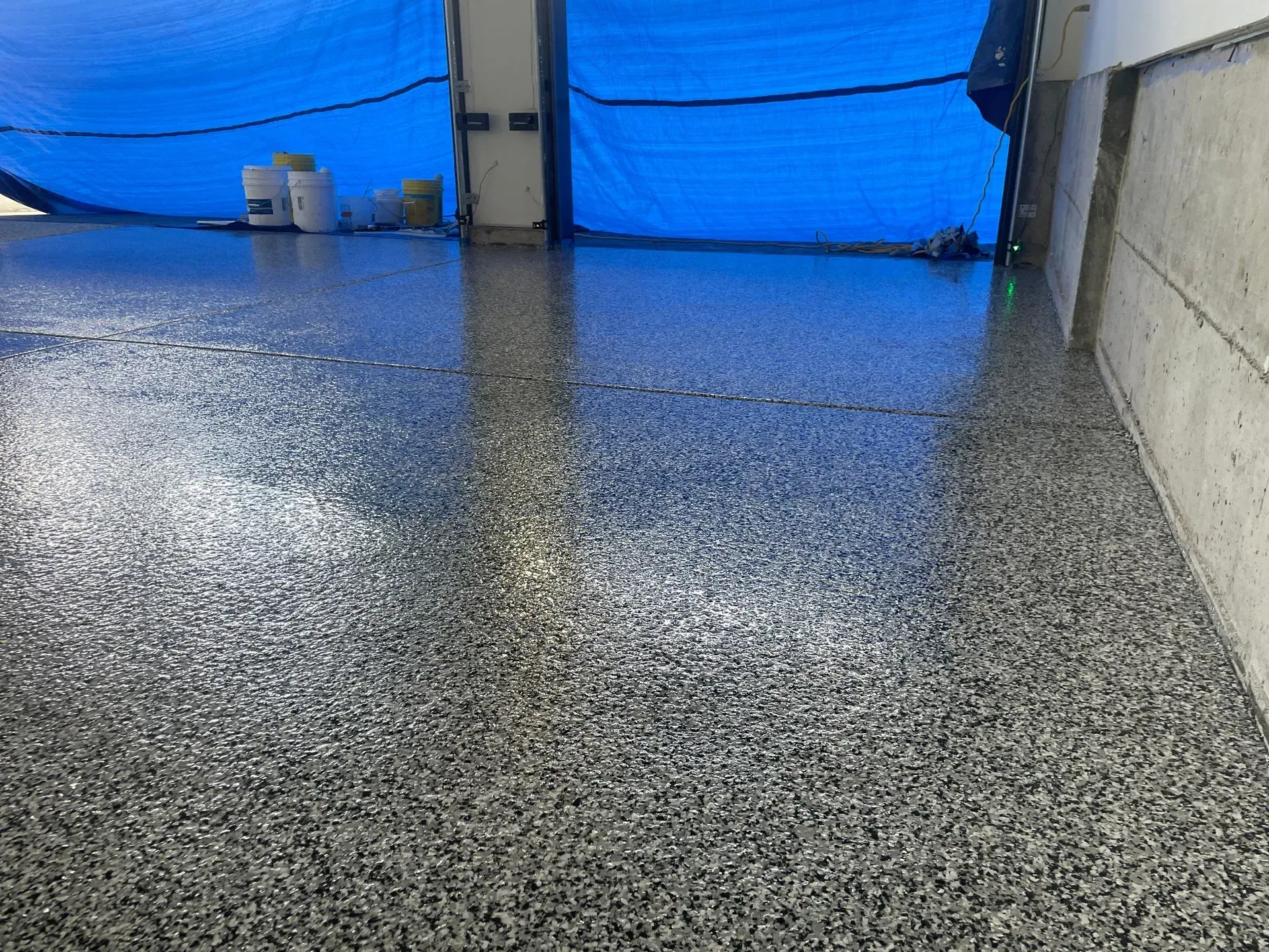 Shiny, speckled gray epoxy floor in an industrial setting, reflecting light. Blue tarp overhead.