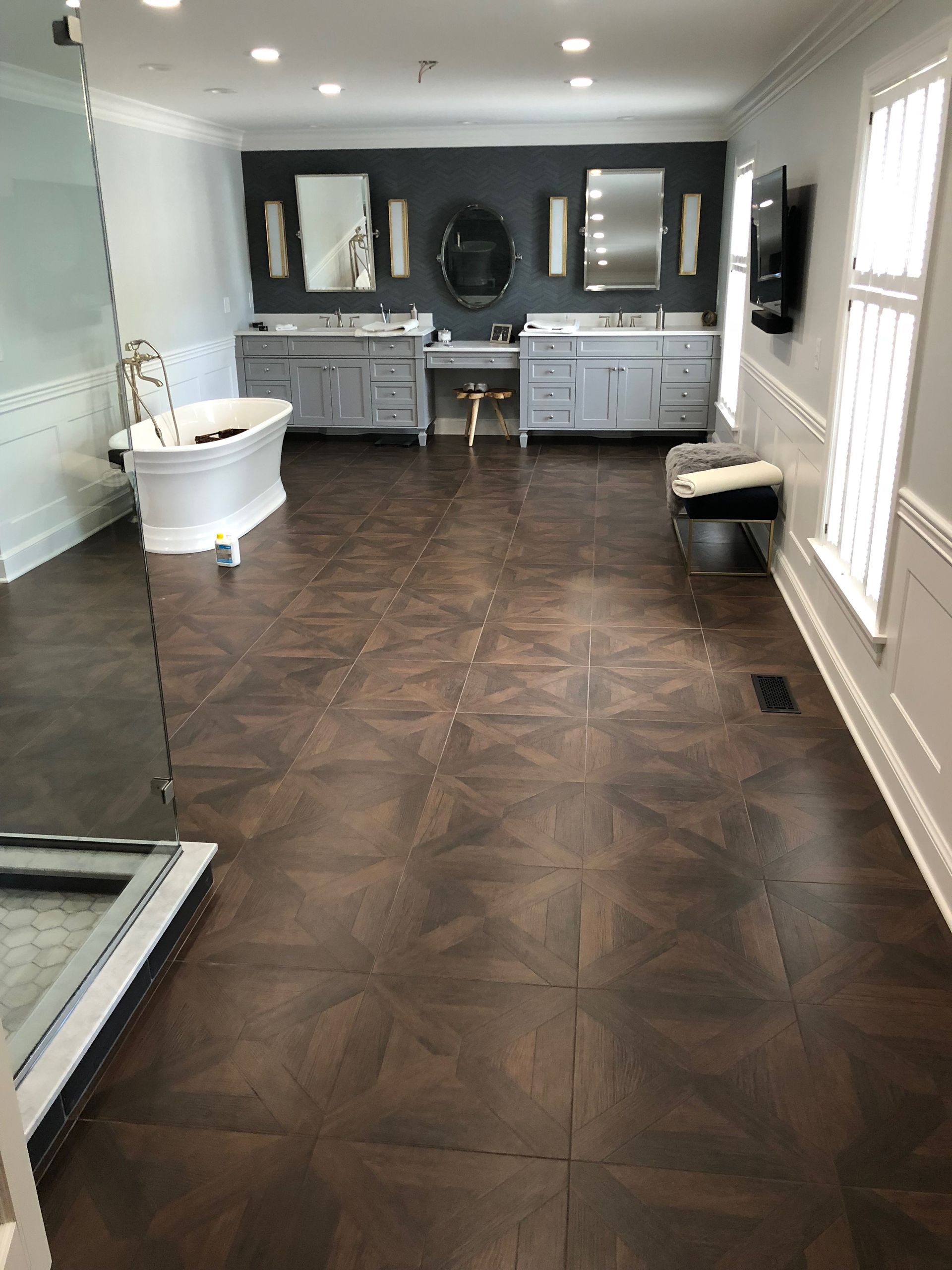 Spacious bathroom with dark parquet floor, two gray vanities, a white tub, and a large glass shower enclosure.