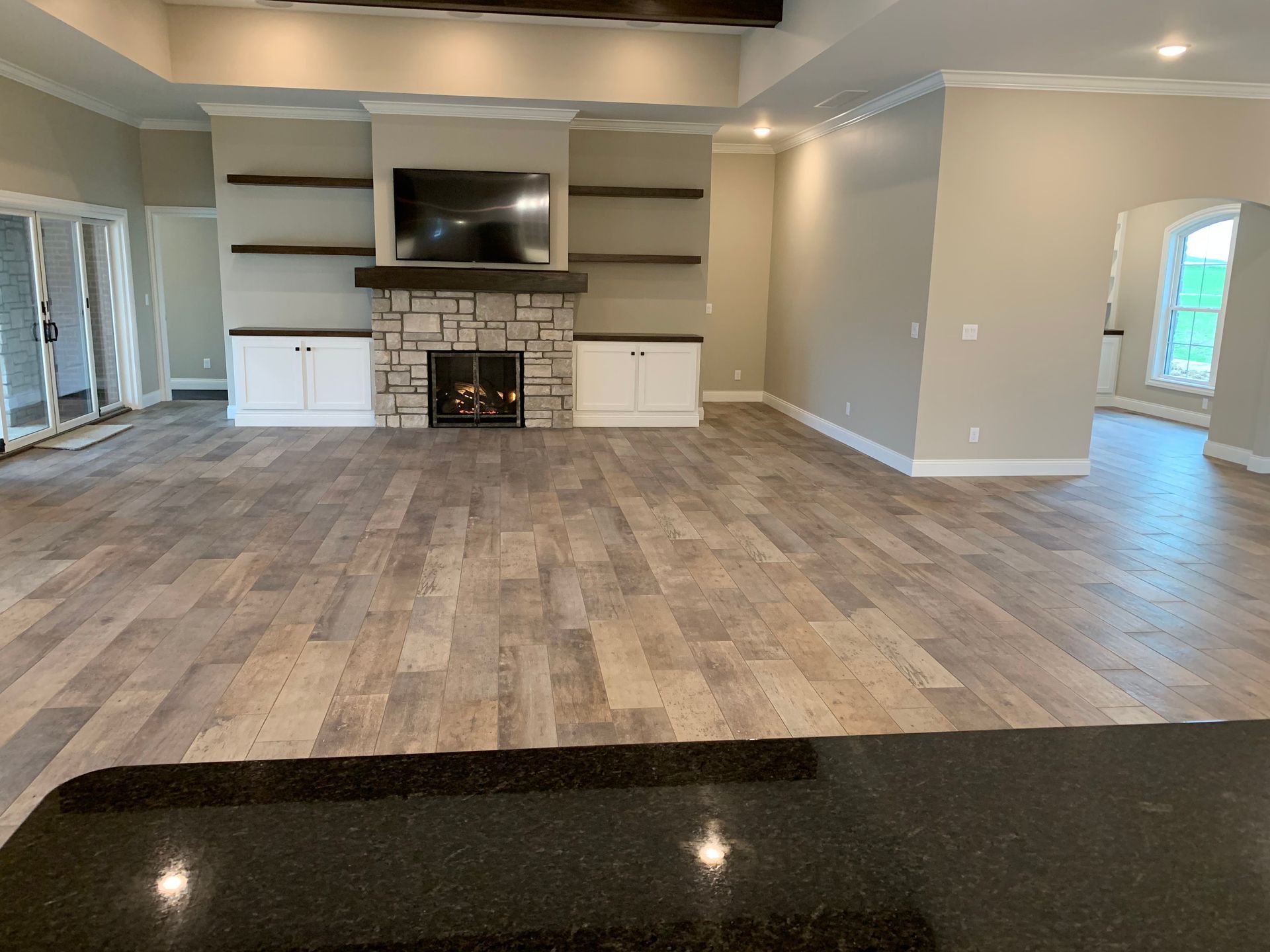 Spacious living room with stone fireplace, built-in shelves, and wood-look tile flooring. Beige walls and white trim.