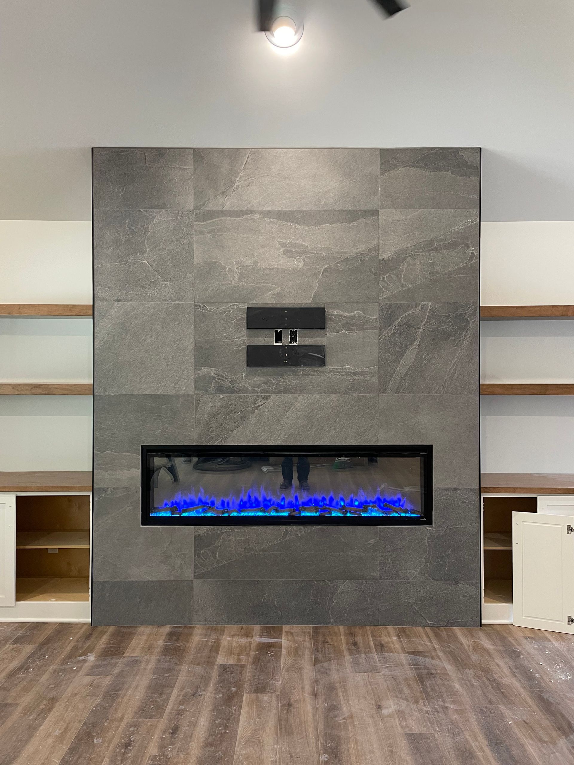Gray tiled fireplace with blue flames, TV mount, and built-in white cabinets/shelves, on wood floor.