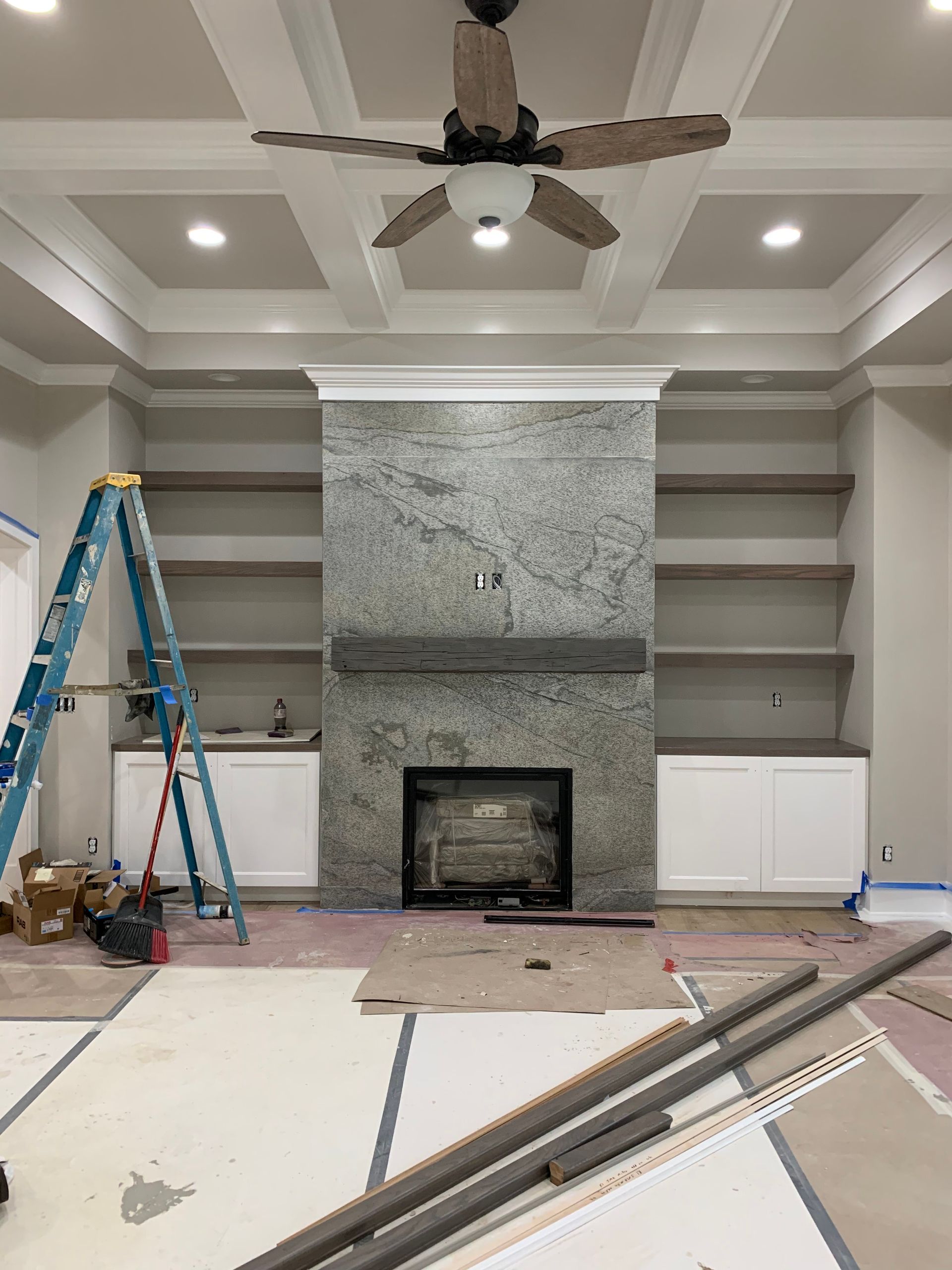 Living room under construction, featuring a fireplace with shelves, cabinets, and a ceiling fan.