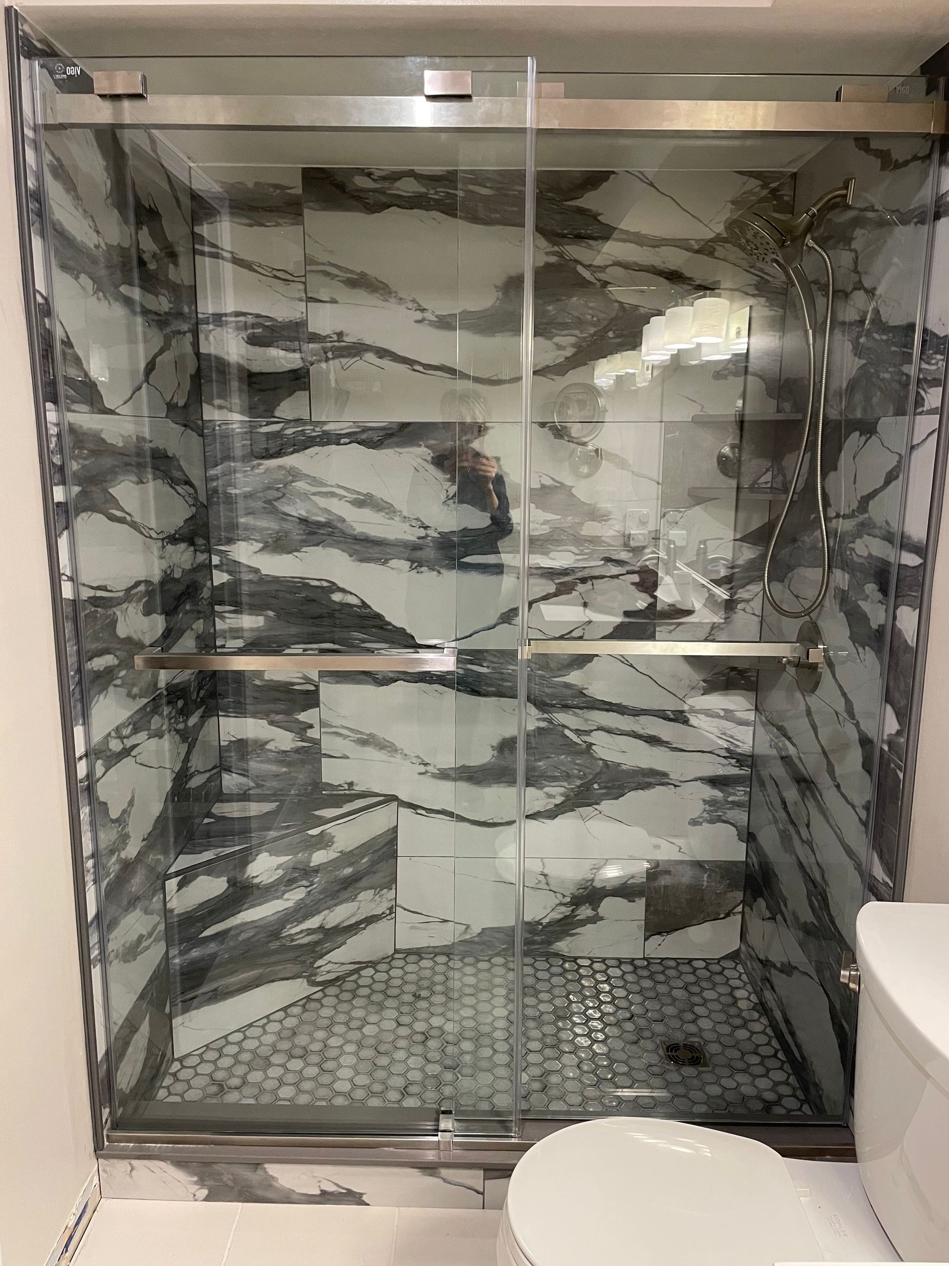 Shower with glass doors and marble-look tile walls. Silver fixtures, small mosaic floor, and a toilet are visible.