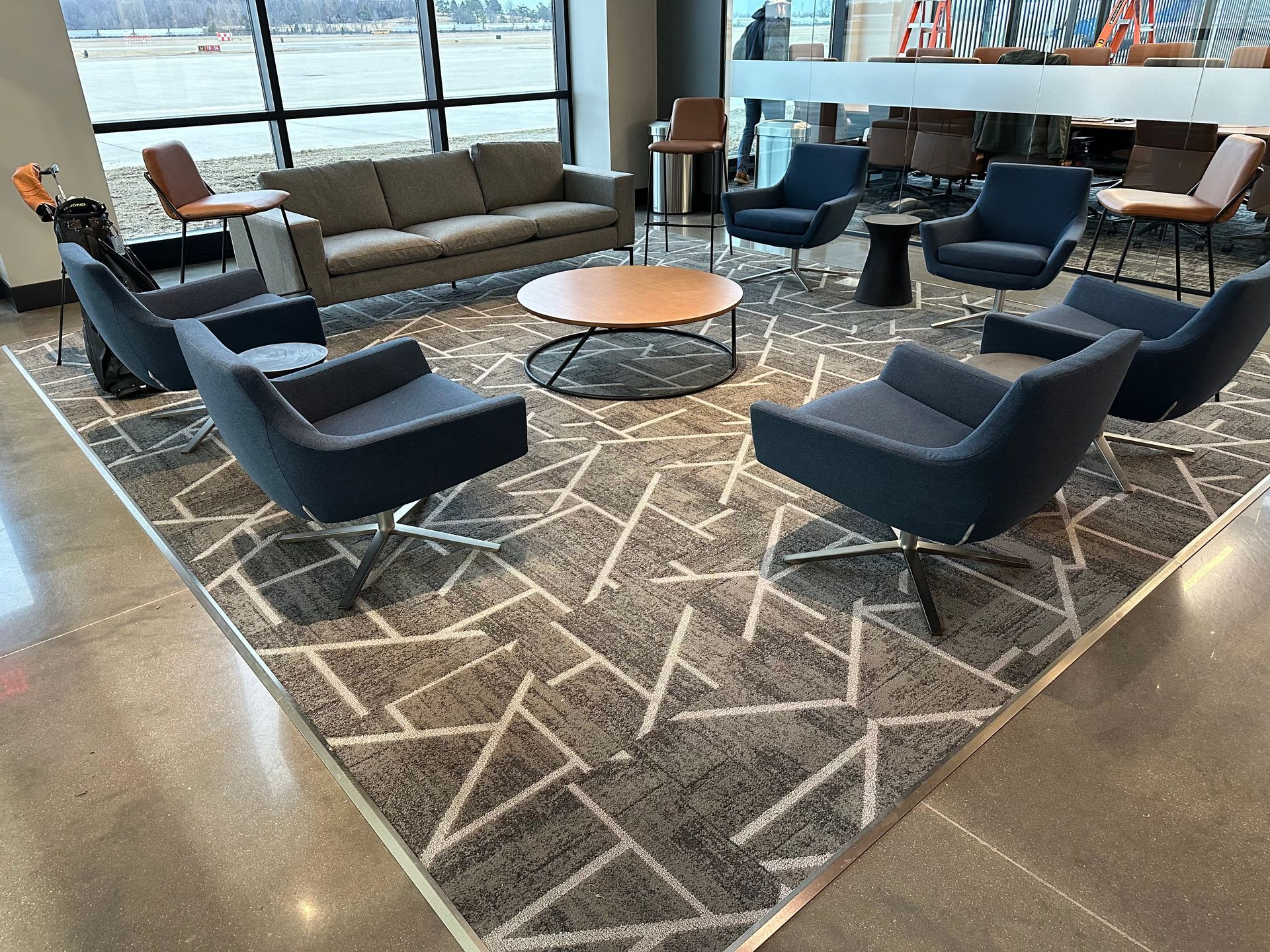 Lounge area with gray sofa, blue chairs, circular table, and patterned rug. Large windows overlook a body of water.
