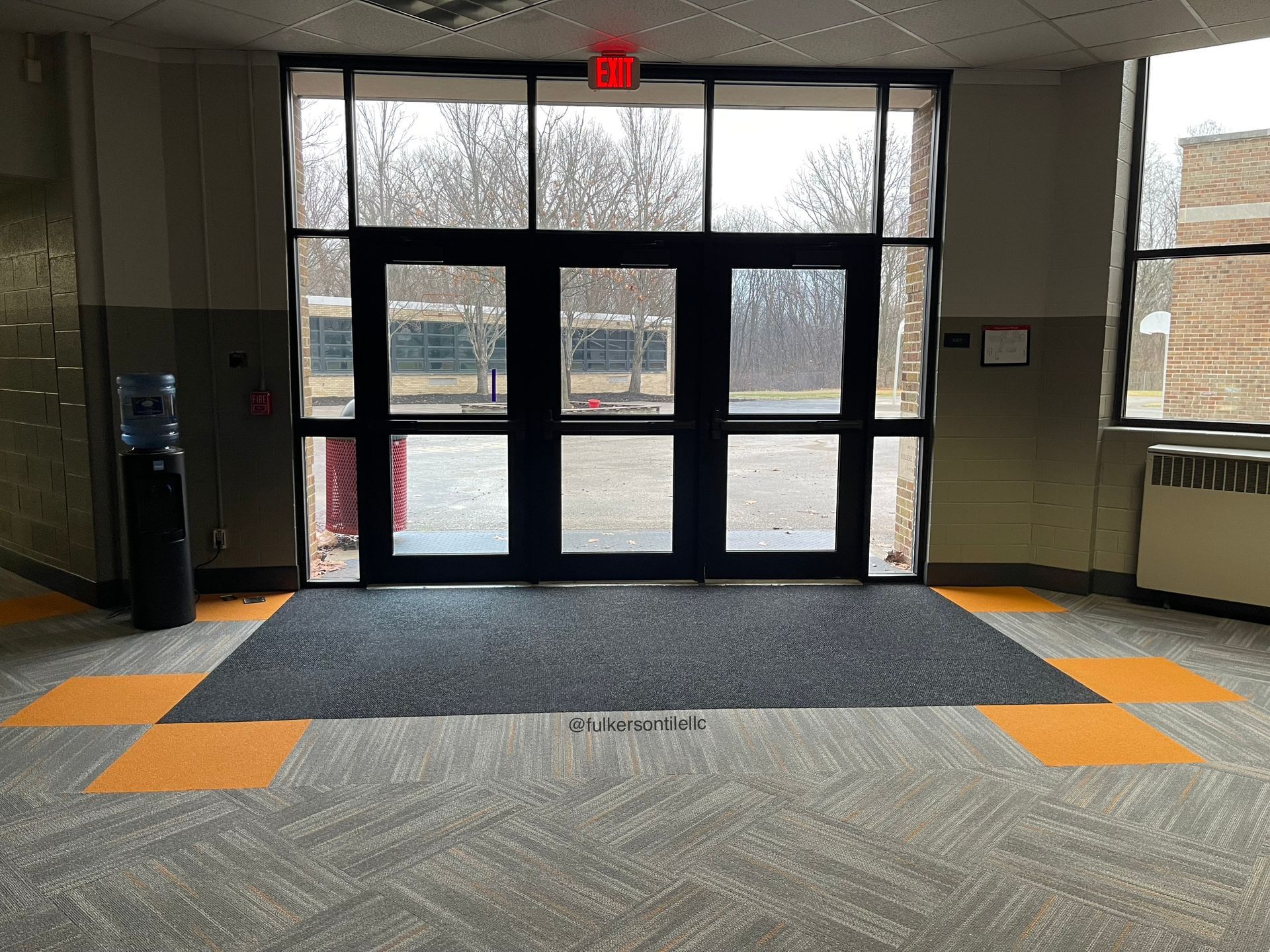 Entrance to a building with glass doors, a doormat, and a water cooler. Gray and orange floor tiles.