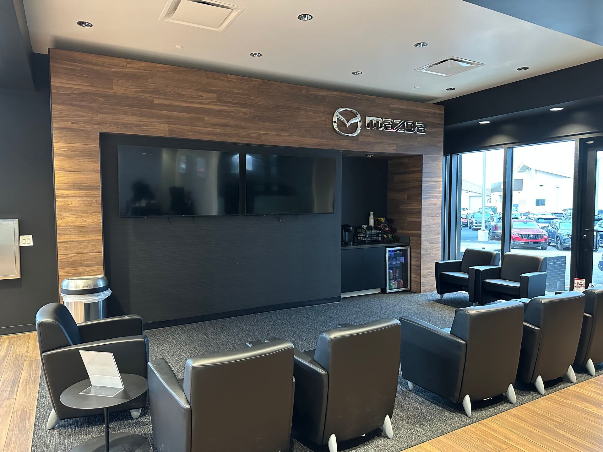 Mazda dealership waiting area: black armchairs, TVs, wood paneling, Mazda logo, coffee station, windows.