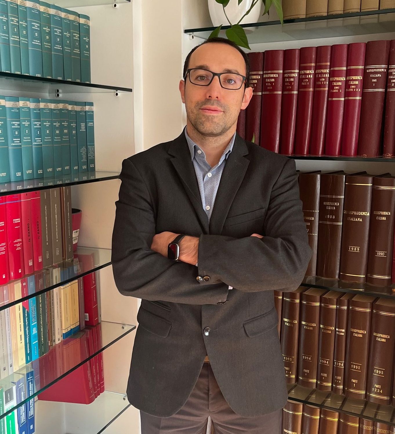 Un uomo con gli occhiali e la giacca, le braccia incrociate, in piedi davanti a scaffali pieni di libri di diritto.