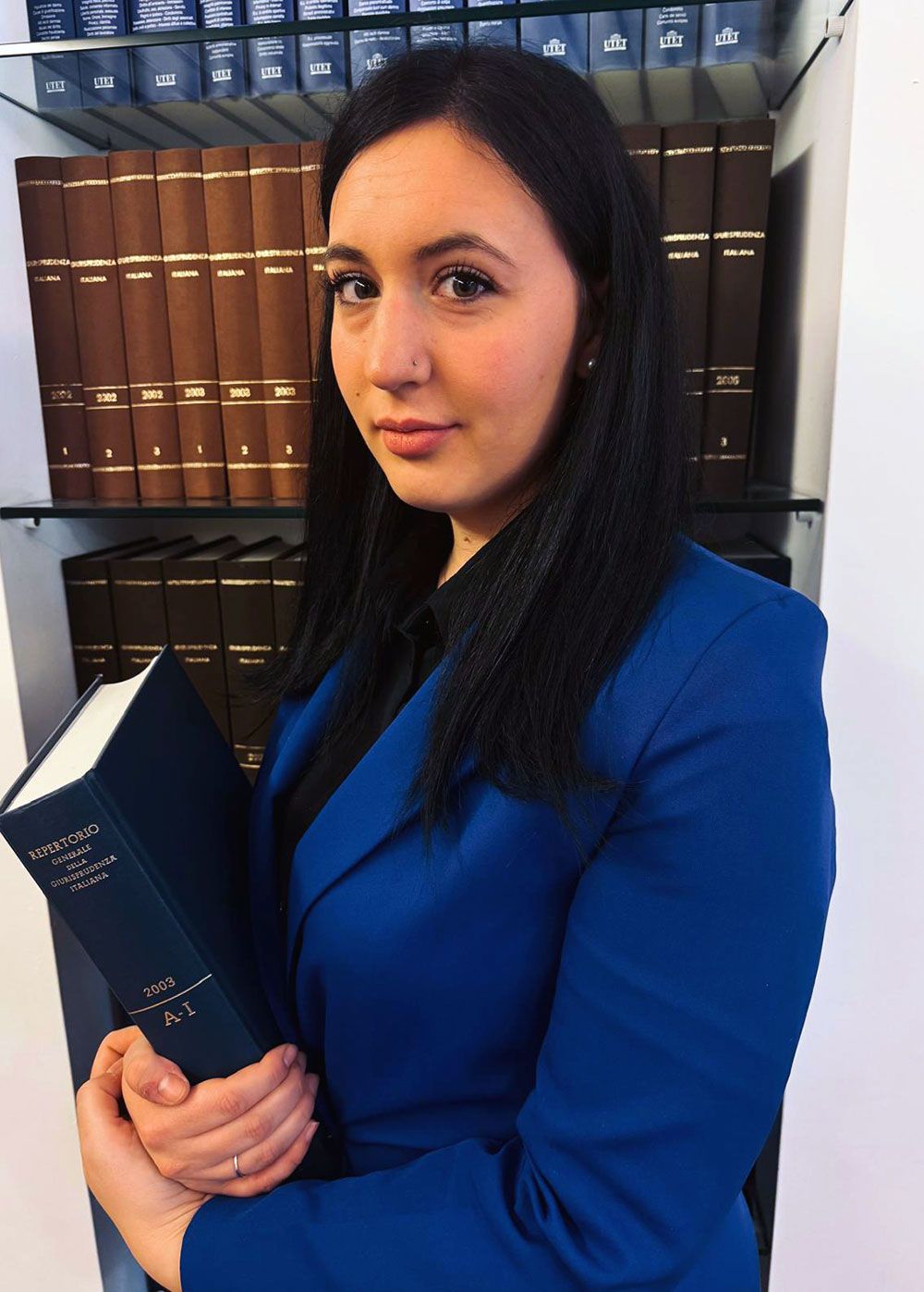 Donna in blazer blu con in mano un libro di giurisprudenza, in piedi davanti agli scaffali.