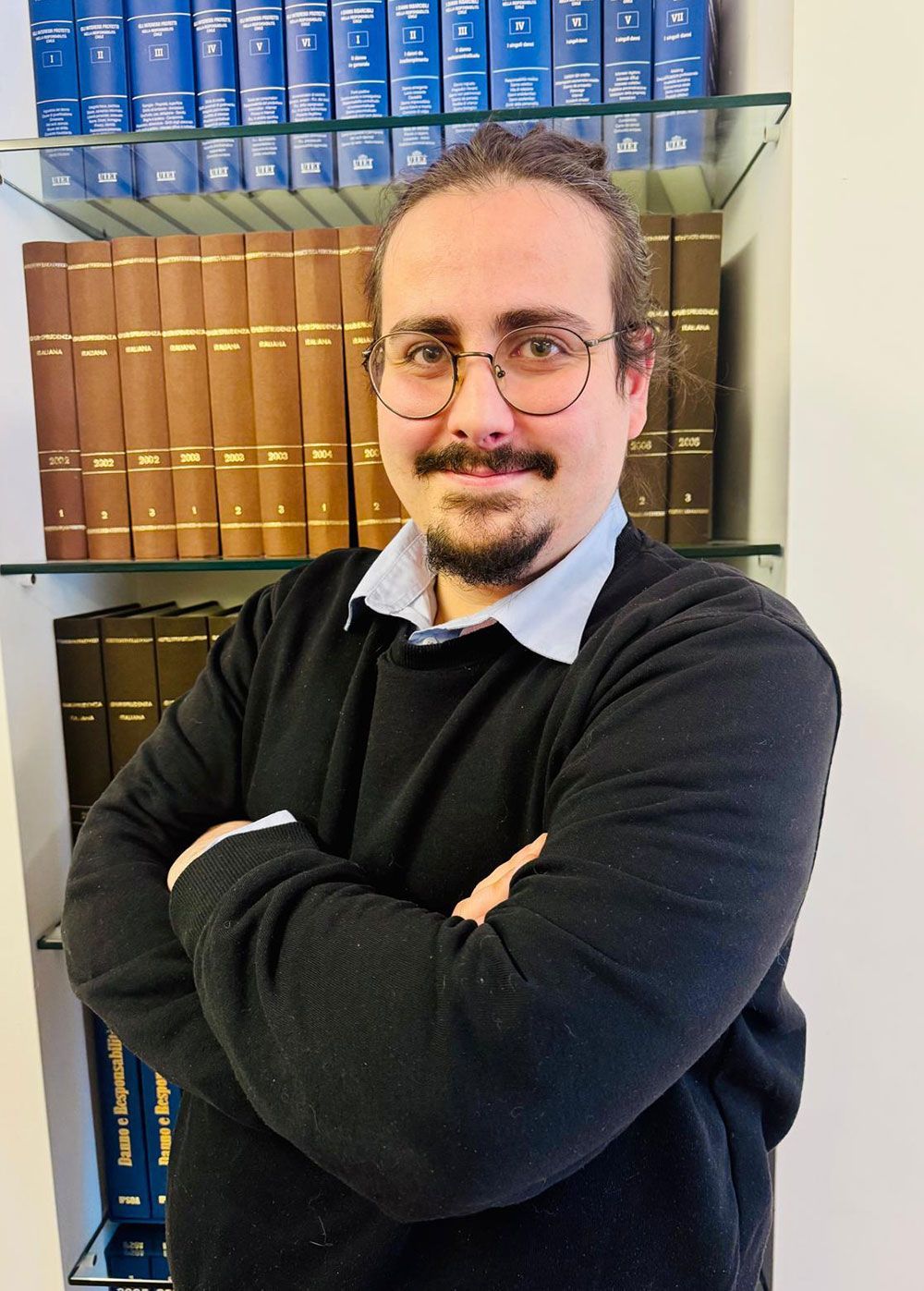 Uomo con occhiali e barba, braccia incrociate, davanti a una libreria piena di libri.