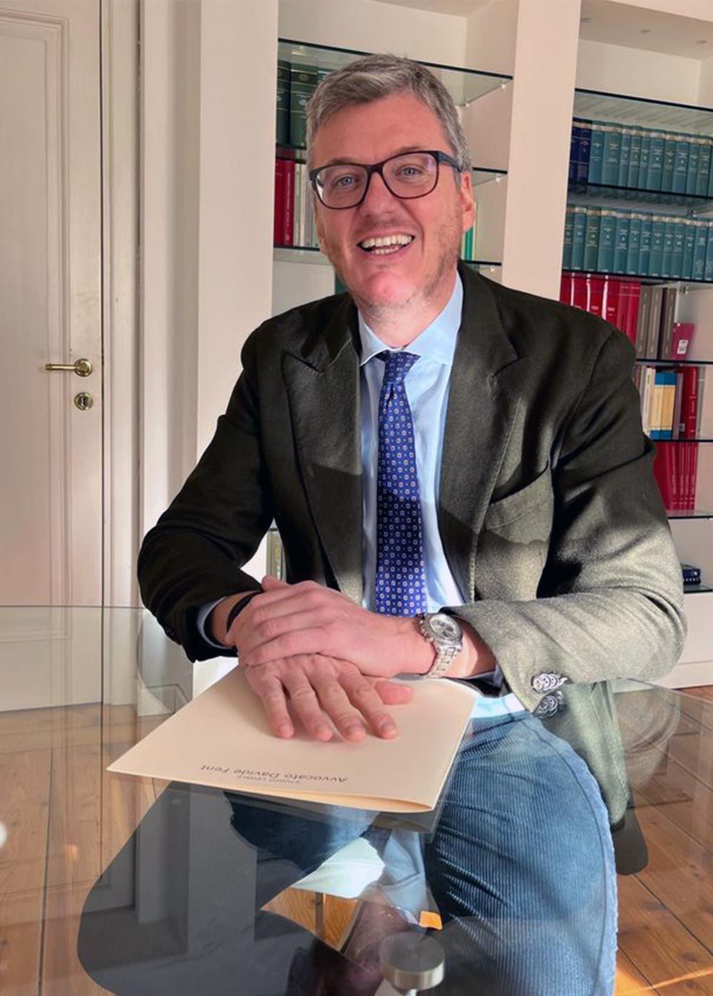 Un uomo con occhiali, blazer e jeans sorride, tenendo in mano un fascicolo, seduto. Sullo sfondo una libreria.