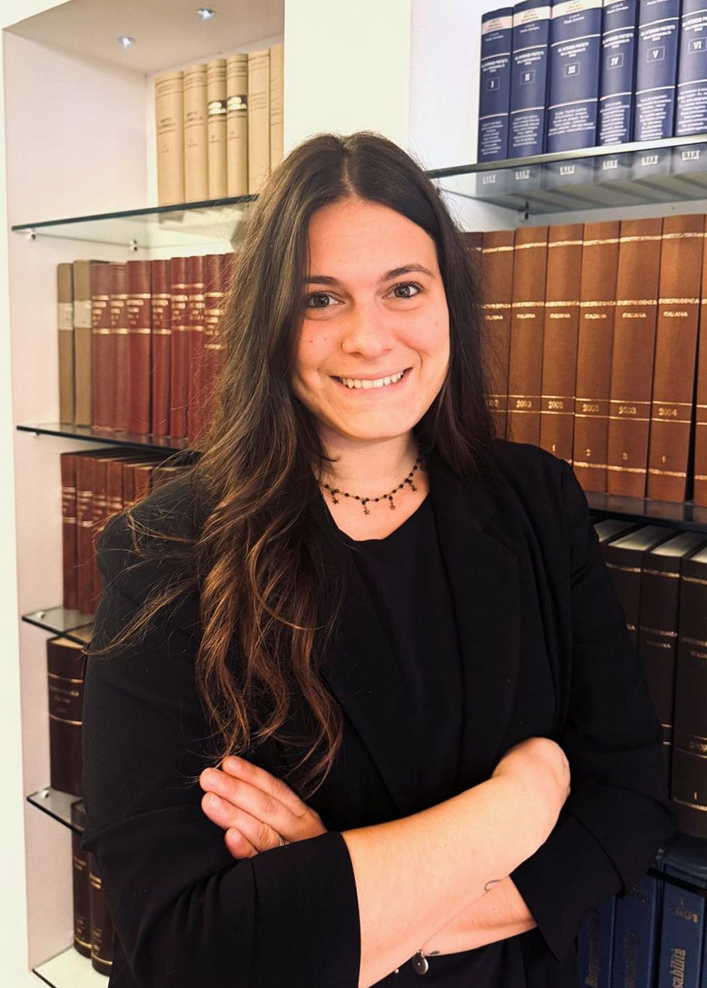 Donna con lunghi capelli castani, braccia incrociate, sorridente. Davanti a una libreria piena di libri.