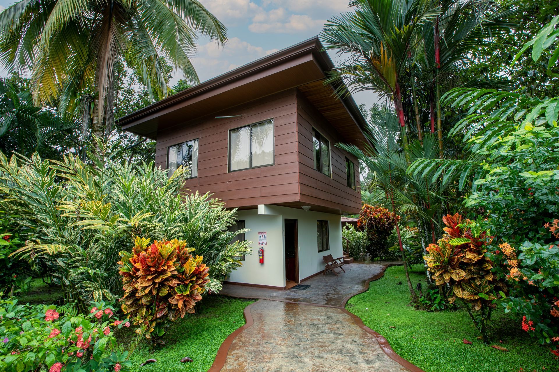 Casa tropical con planta superior de madera y sendero de piedra, rodeada de exuberantes plantas verdes y coloridos arbustos.