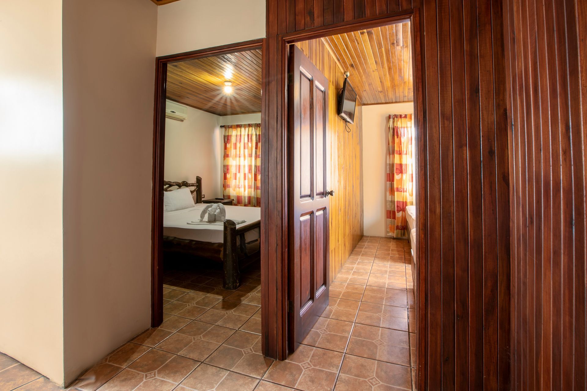 Pasillo revestido de madera con suelo de baldosas que conduce a un dormitorio con cama y cortinas estampadas.
