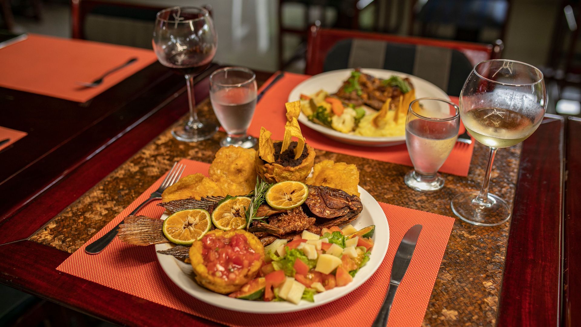 Dos platos servidos en una mesa de restaurante con copas de vino y manteles individuales rojos.