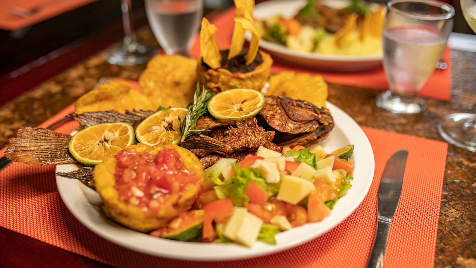 Plato de verduras y frutas variadas a la parrilla sobre una mesa con mantel individual naranja y cristalería.