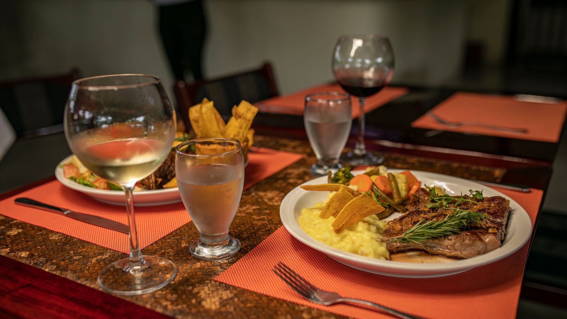 Mesa de cena con copas de vino, un cóctel y pescado y verduras emplatados sobre manteles individuales rojos.