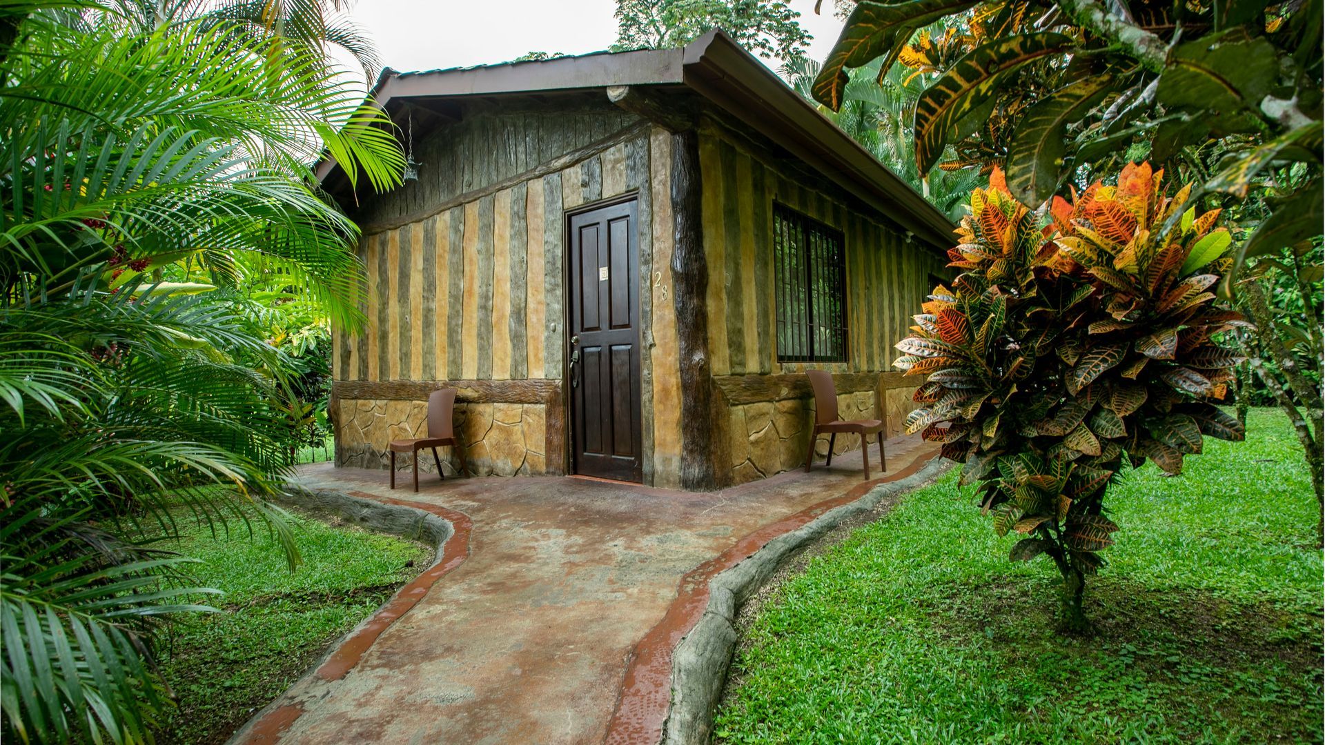 Pequeña cabaña rústica con un sendero curvo de piedra, rodeada de exuberante vegetación tropical y plantas coloridas.
