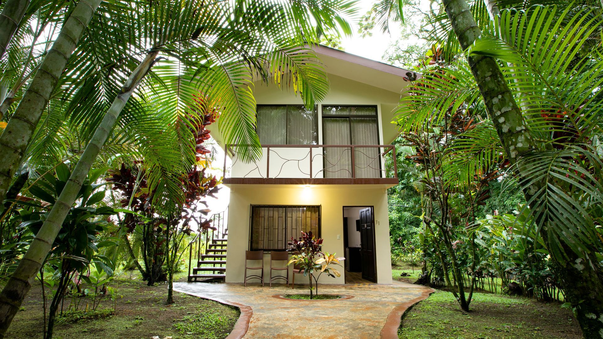 Casa de dos plantas con balcón, rodeada de exuberante vegetación tropical y un sendero curvo de piedra.