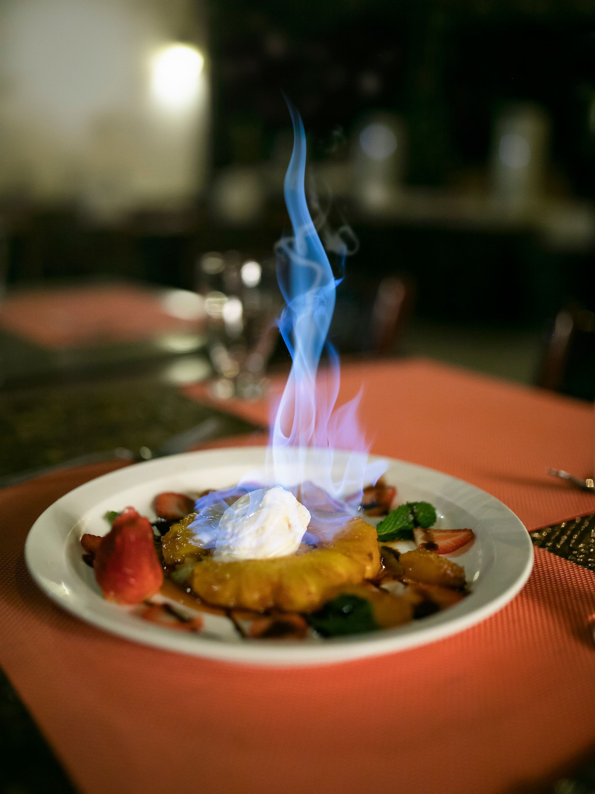 Postre flameado en un plato blanco sobre una mesa de restaurante iluminada con velas y cubierta con un mantel rojo.