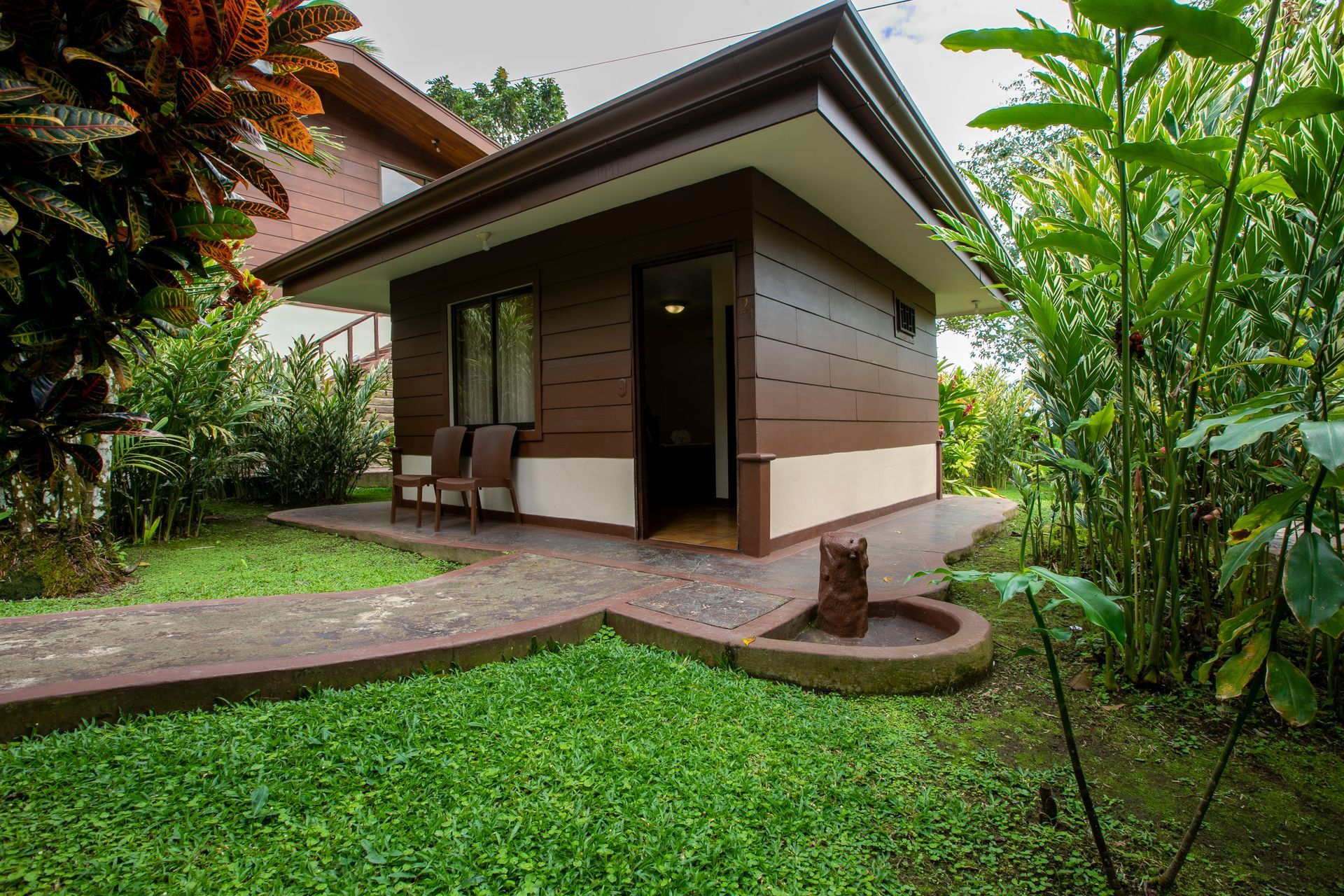 Pequeña casa moderna con porche, rodeada de un exuberante jardín verde y plantas tropicales.