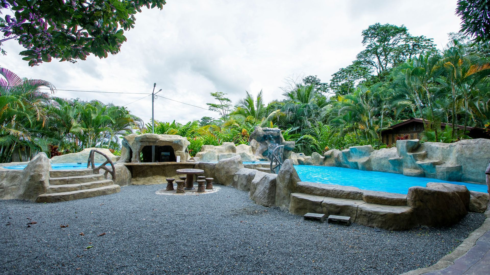 Zona de spa al aire libre con jacuzzis de piedra, piscinas azules y grava rodeada de exuberante vegetación.