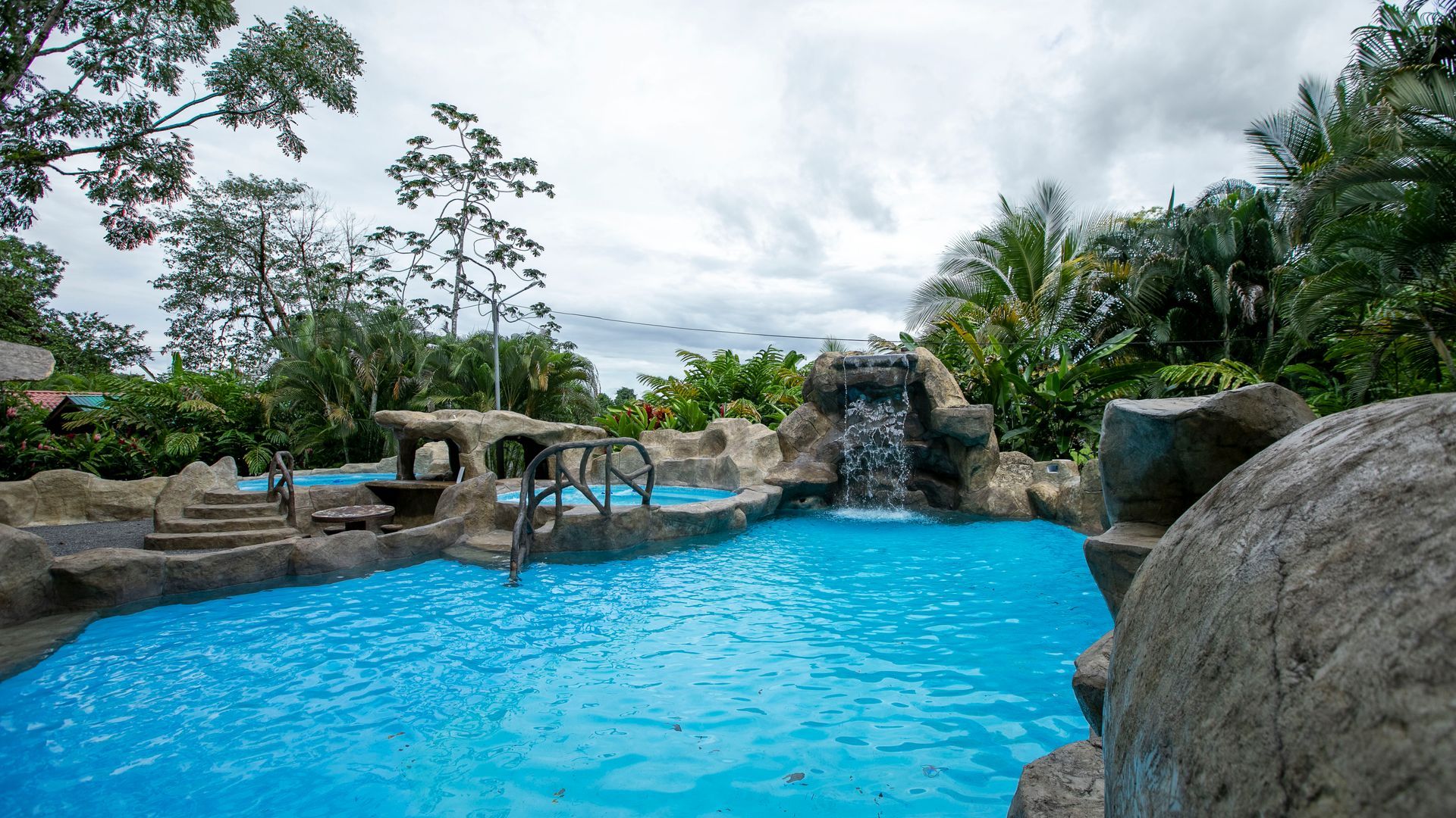 Piscina de un complejo turístico tropical con agua azul brillante, rocas y palmeras bajo un cielo nublado.