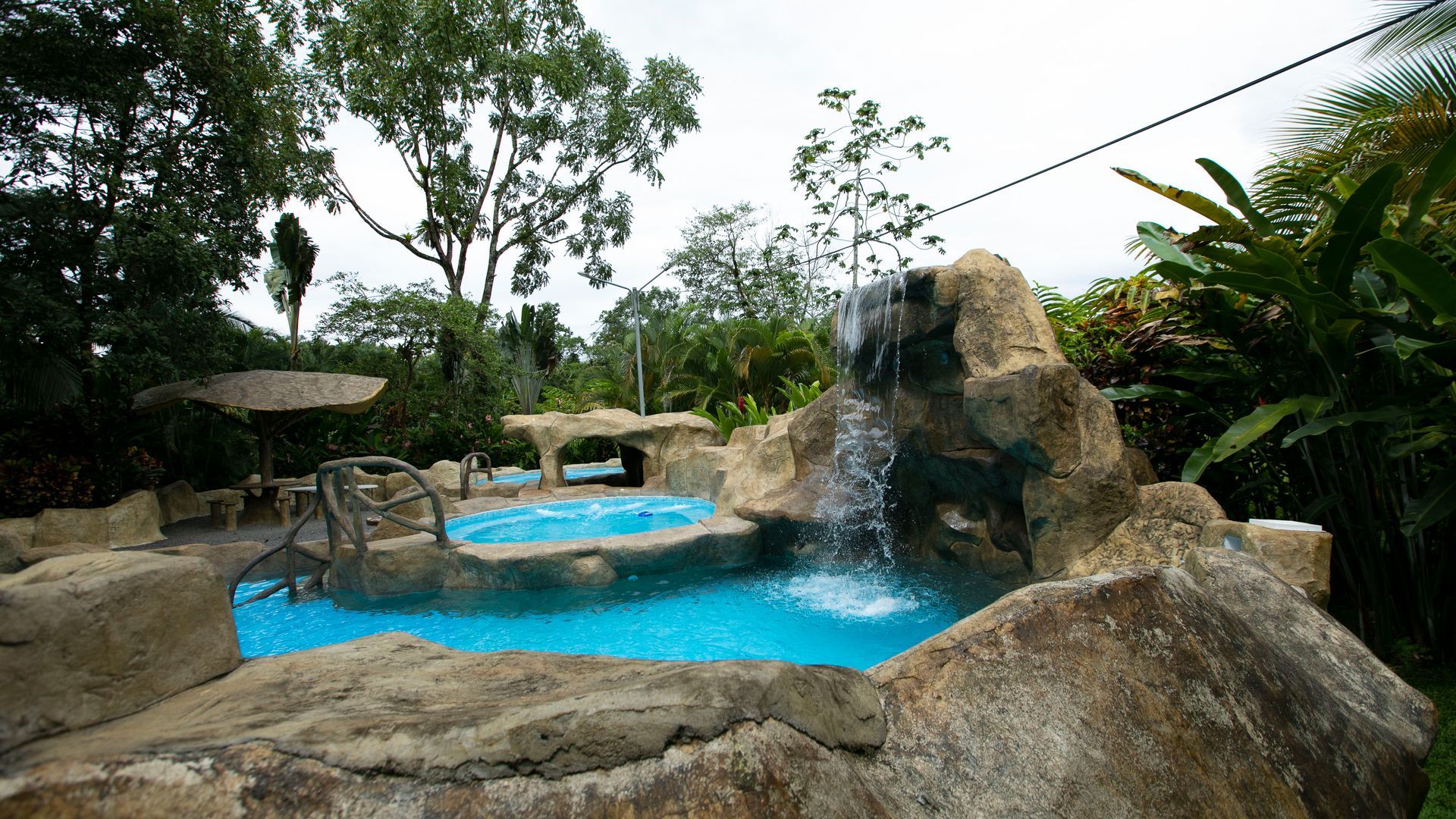 Piscina exterior rocosa con agua azul brillante y una pequeña cascada rodeada de vegetación tropical.