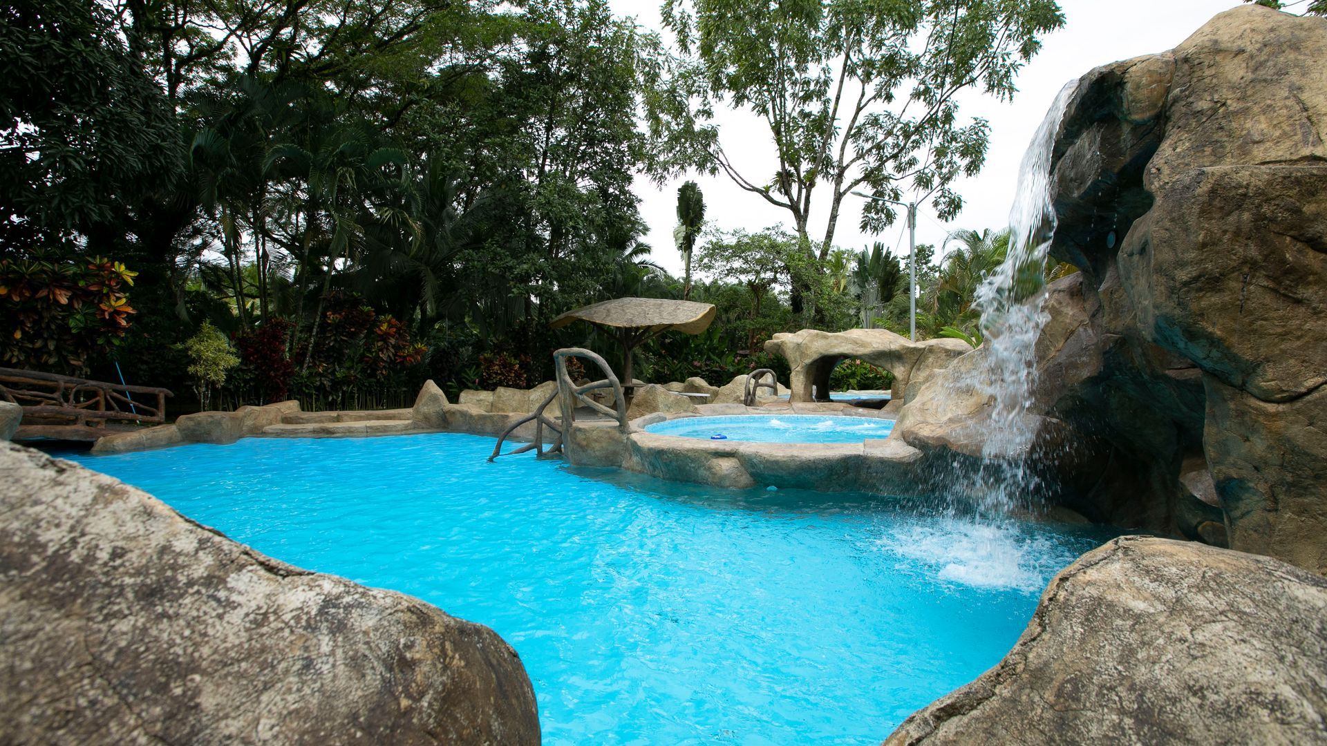 Piscina azul estilo resort con cascadas y formaciones rocosas rodeadas de exuberantes árboles.