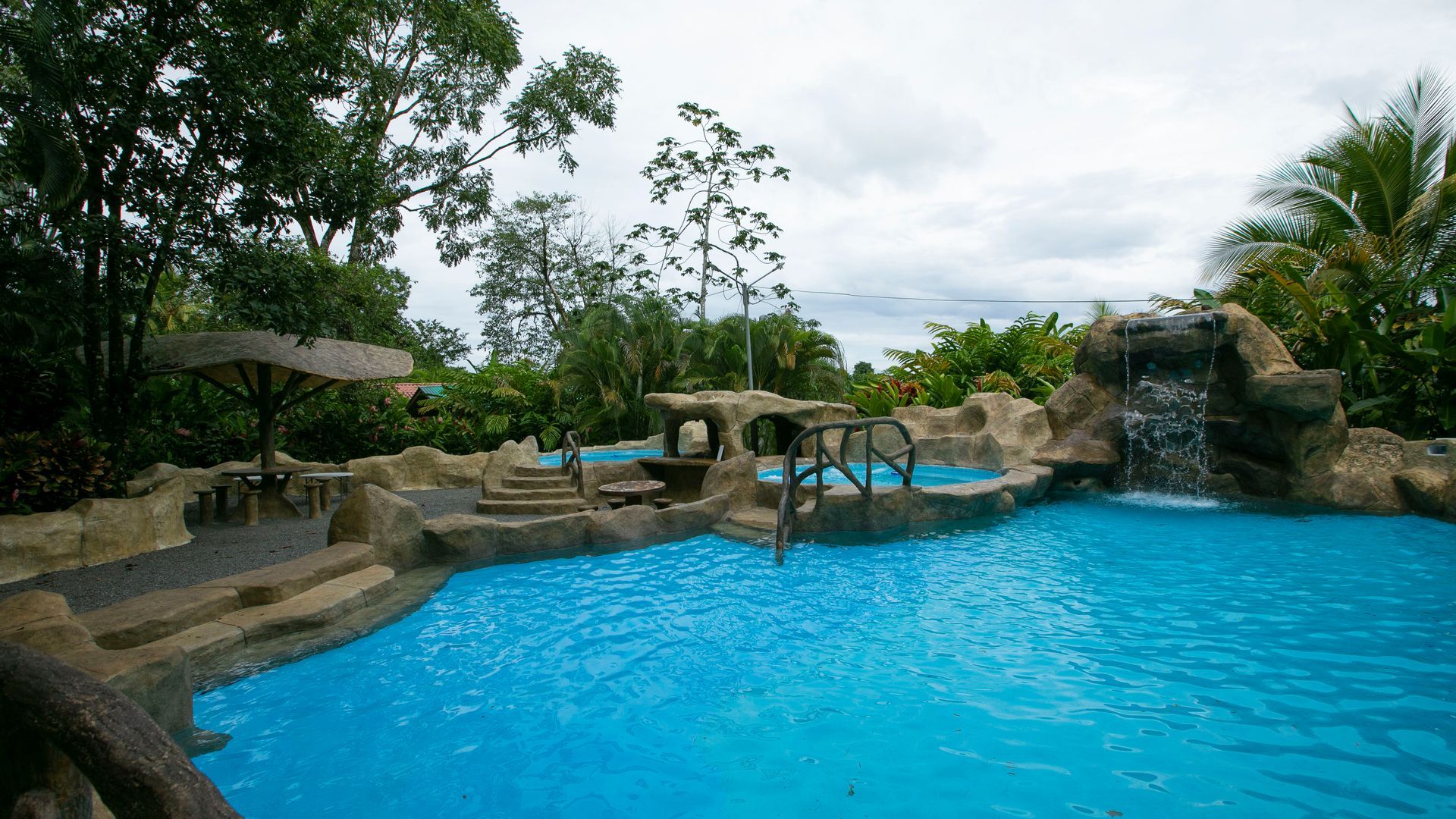 Piscina tropical en un complejo turístico con agua azul brillante, rocas, árboles y un pequeño puente bajo un cielo nublado.