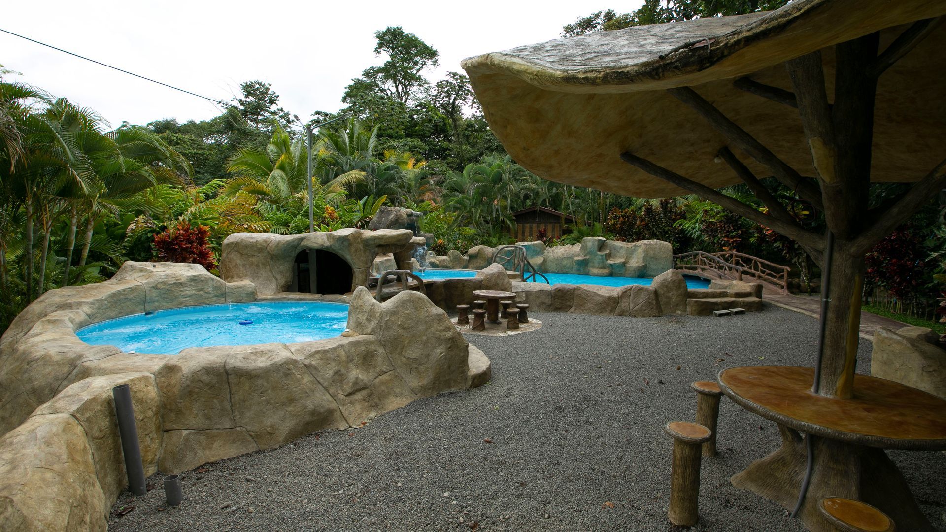 Zona de piscinas de estilo tropical con jacuzzis azules, cascadas de piedra, senderos de grava y una sombrilla de paja.