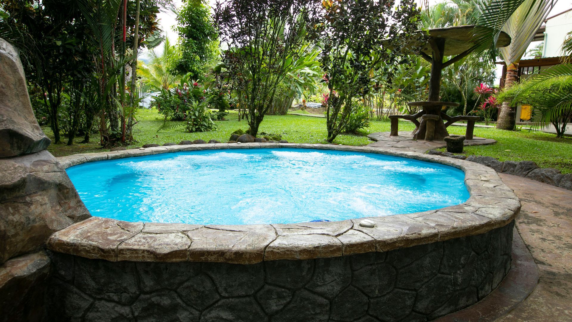 Piscina redonda revestida de piedra, llena de agua azul brillante, en un entorno de jardín exuberante.