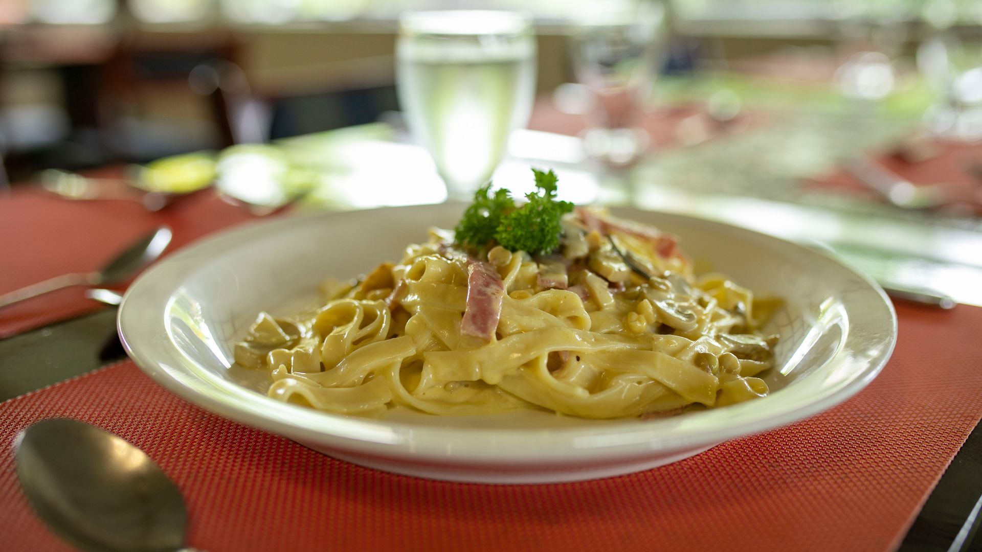 Pasta en un tazón blanco sobre un mantel individual rojo en la mesa de un restaurante, cubierta con hierbas y jamón.