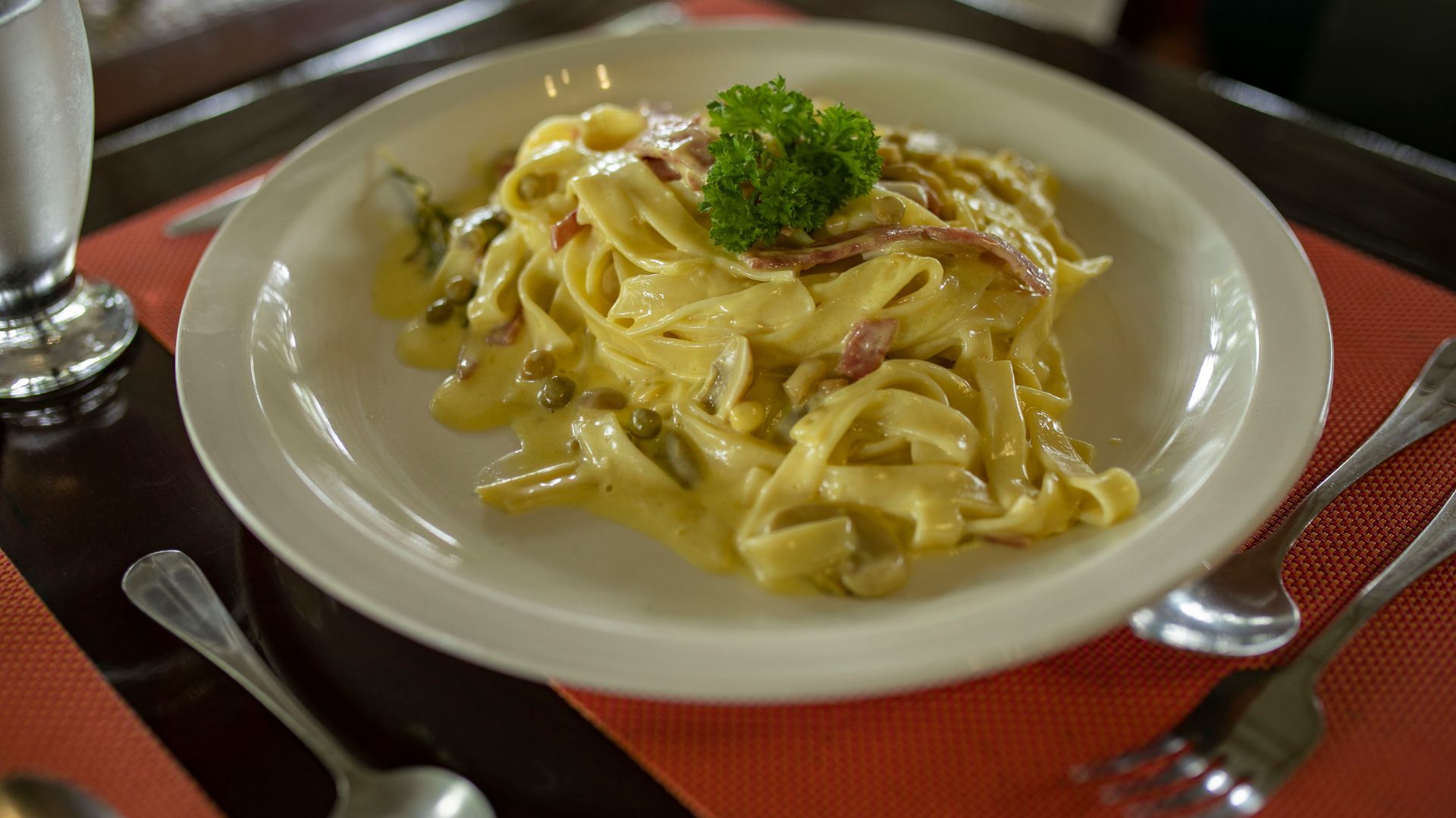 Plato de pasta cremosa con guarnición de perejil sobre un mantel individual rojo, con cubiertos al lado.