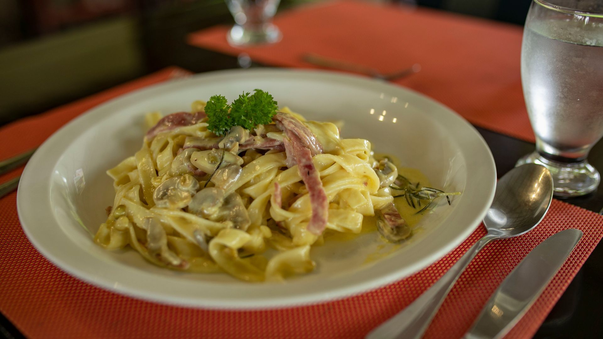 Plato de pasta cremosa con tocino y perejil sobre un mantel individual rojo, junto a un vaso y cubiertos.