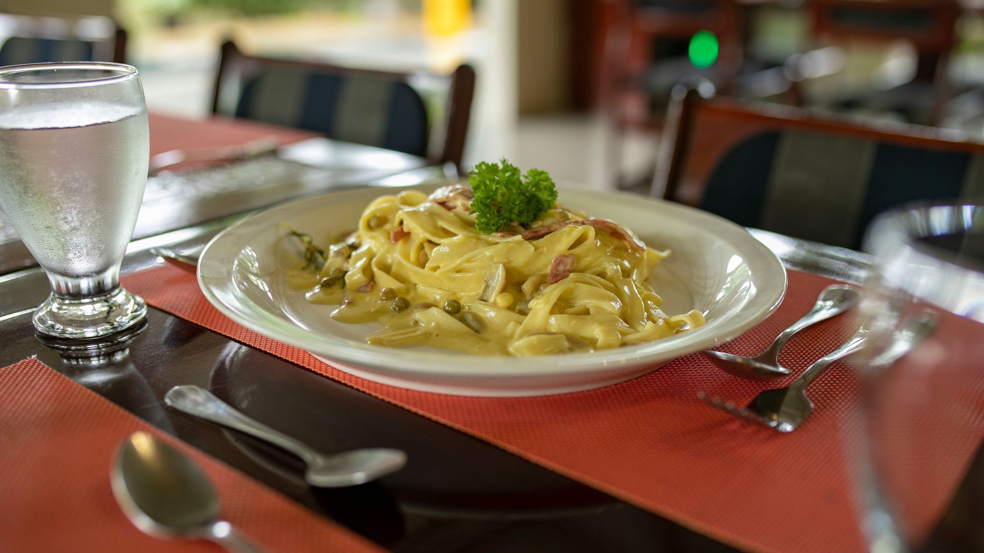 Un plato de pasta cremosa con perejil sobre una mesa roja de restaurante, junto a un vaso de agua.