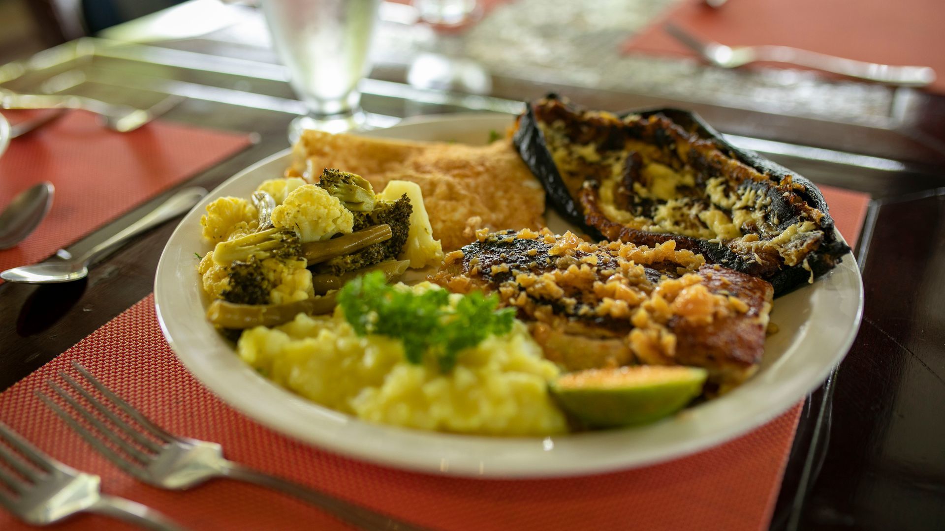 Plato de comida variada sobre un mantel individual rojo en una mesa de comedor, con una copa de vino y cubiertos cerca.