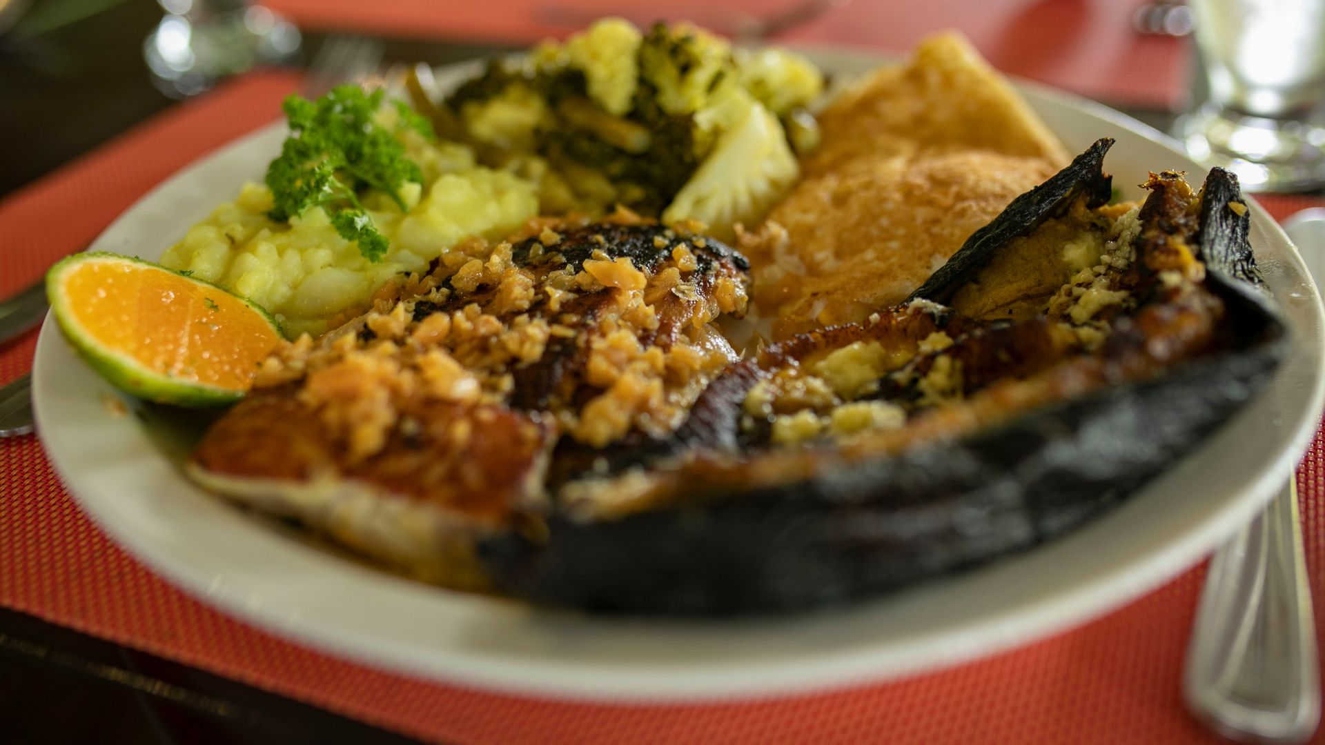Plato de comida variada con brócoli, rodaja de naranja y carne a la parrilla sobre un mantel rojo.