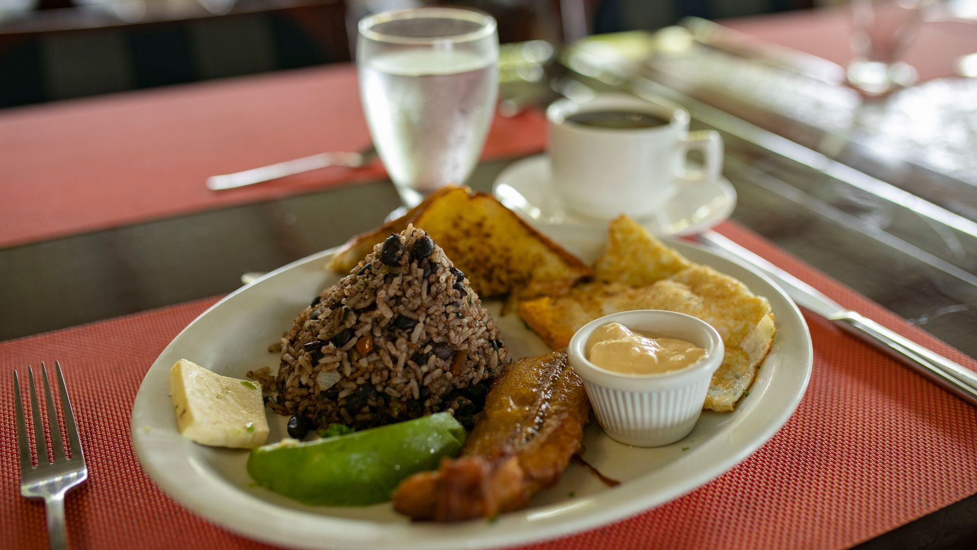 Plato con una variedad de platos tropicales sobre un mantel individual rojo, con un vaso de agua y una taza de café al fondo.