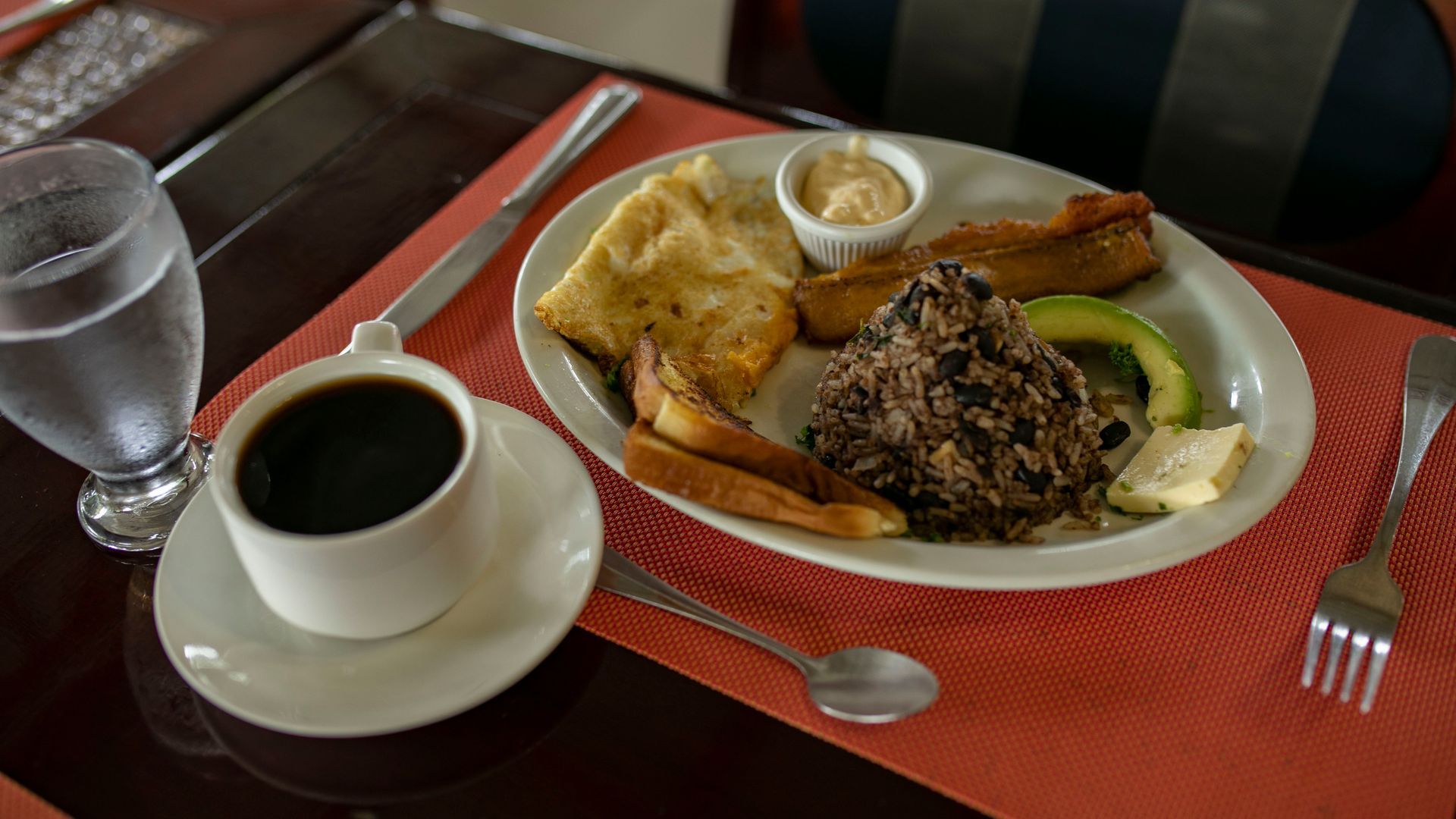 Plato de desayuno con tostadas y patatas fritas junto a una taza de café sobre un mantel individual rojo.