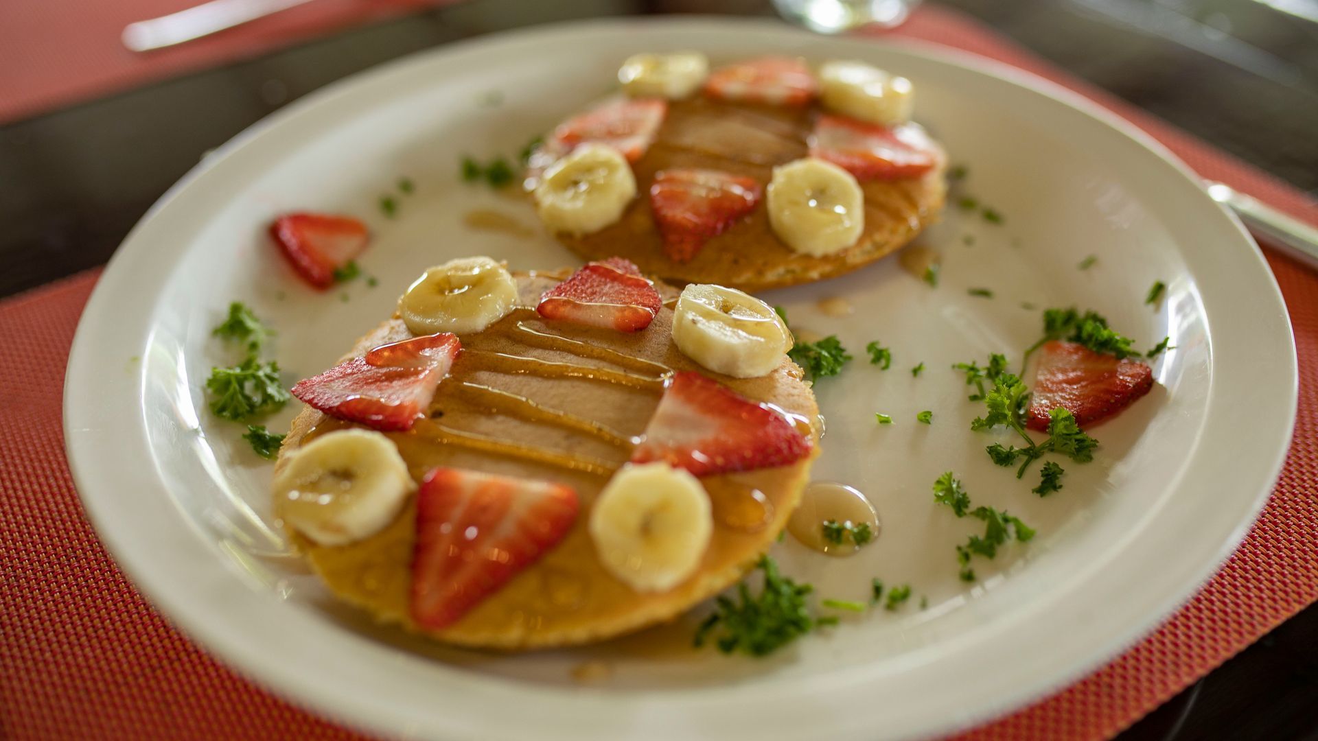 Postre emplatado con fruta en rodajas y crema en un plato blanco, adornado con hierbas sobre un mantel individual rojo.
