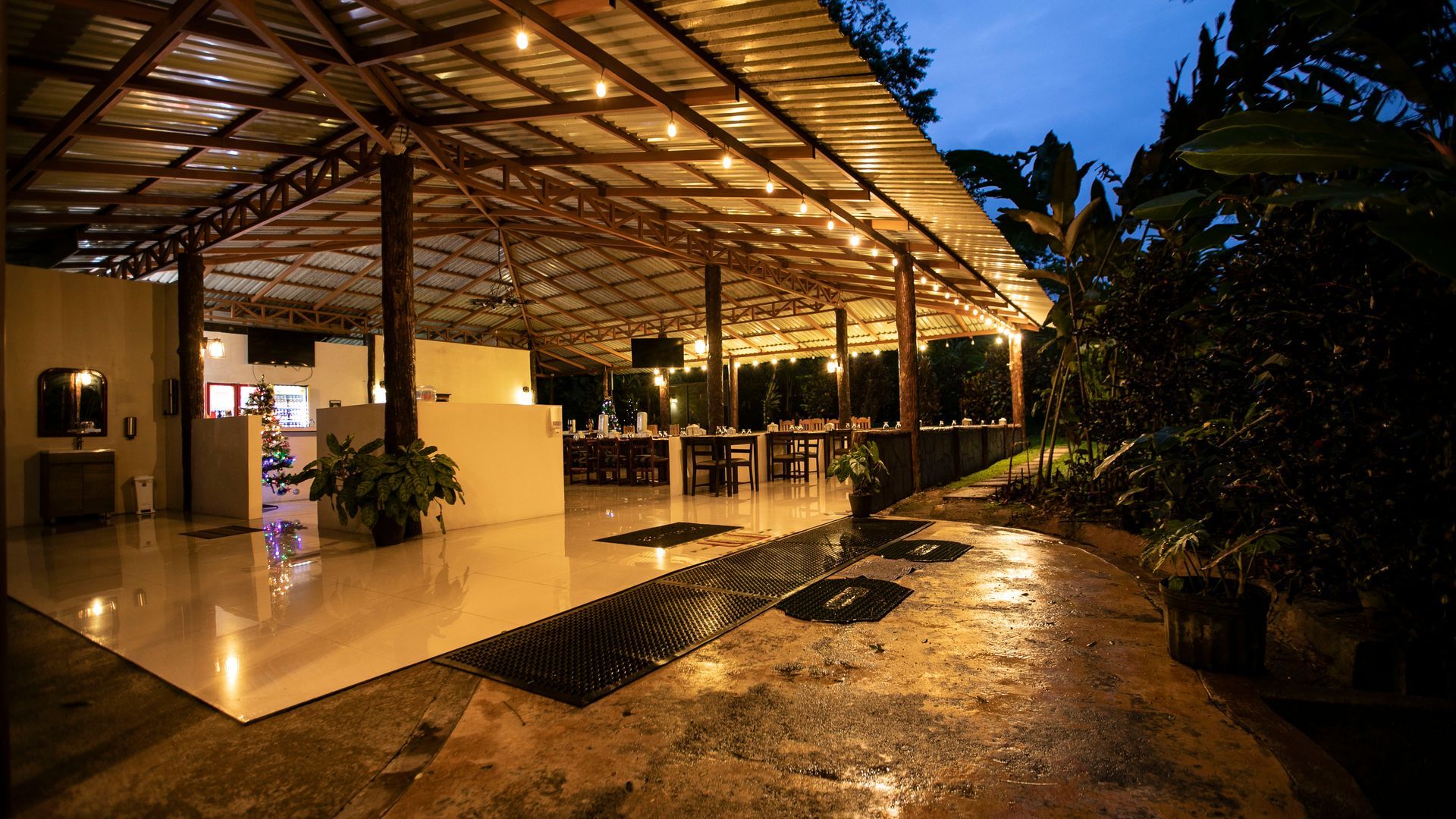 Terraza de restaurante al aire libre al atardecer con luces cálidas, suelo de piedra mojada y plantas en macetas.